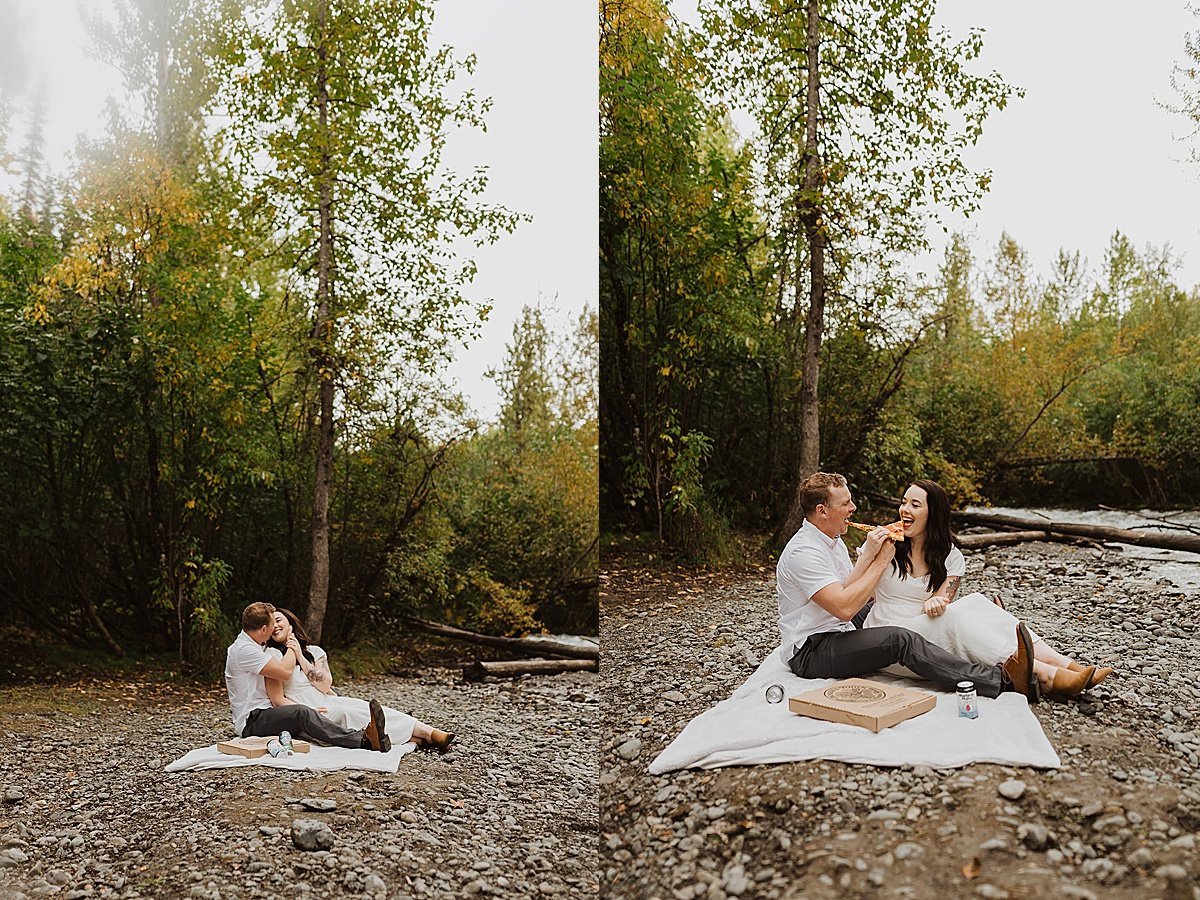  Couple lounges happily on picnic blanket by river in woodsy engagement shoot 