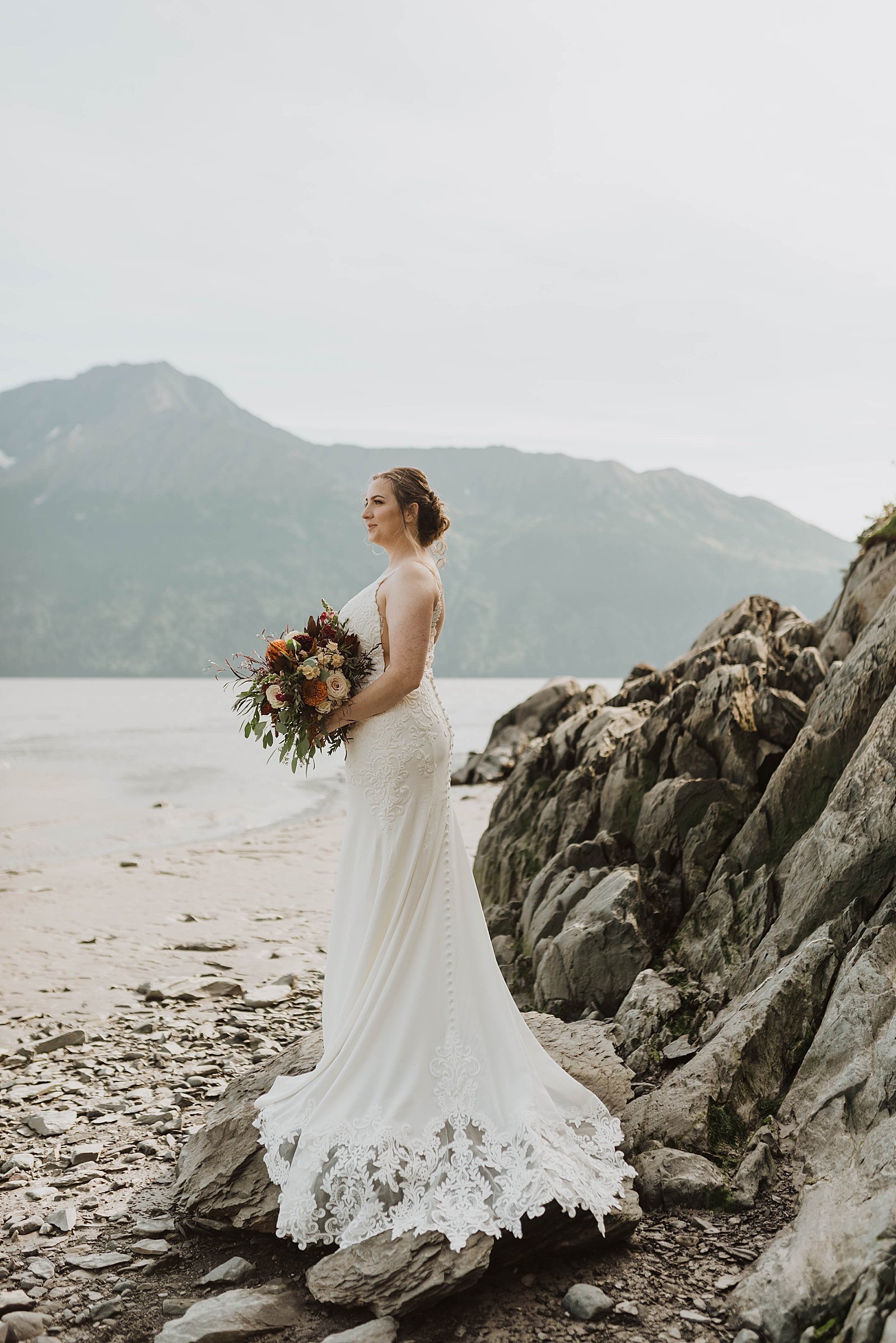  Alaska bride standing on a rock in front of the water with mountains in the background 