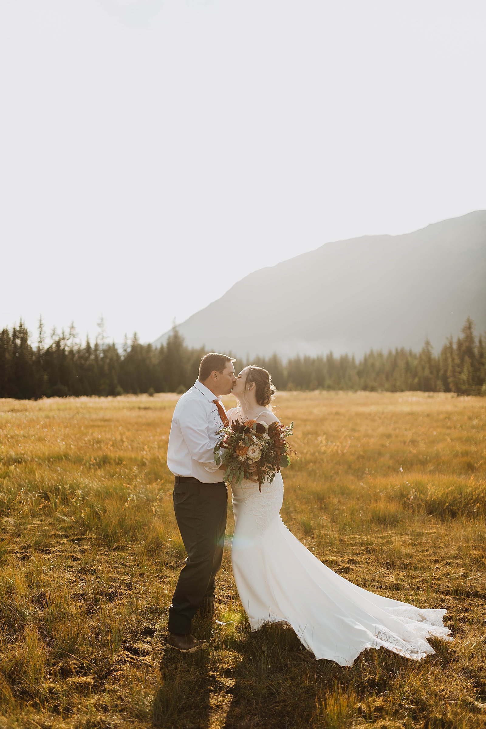  Newlyweds embracing in a field with Girdwood mountains behind them 