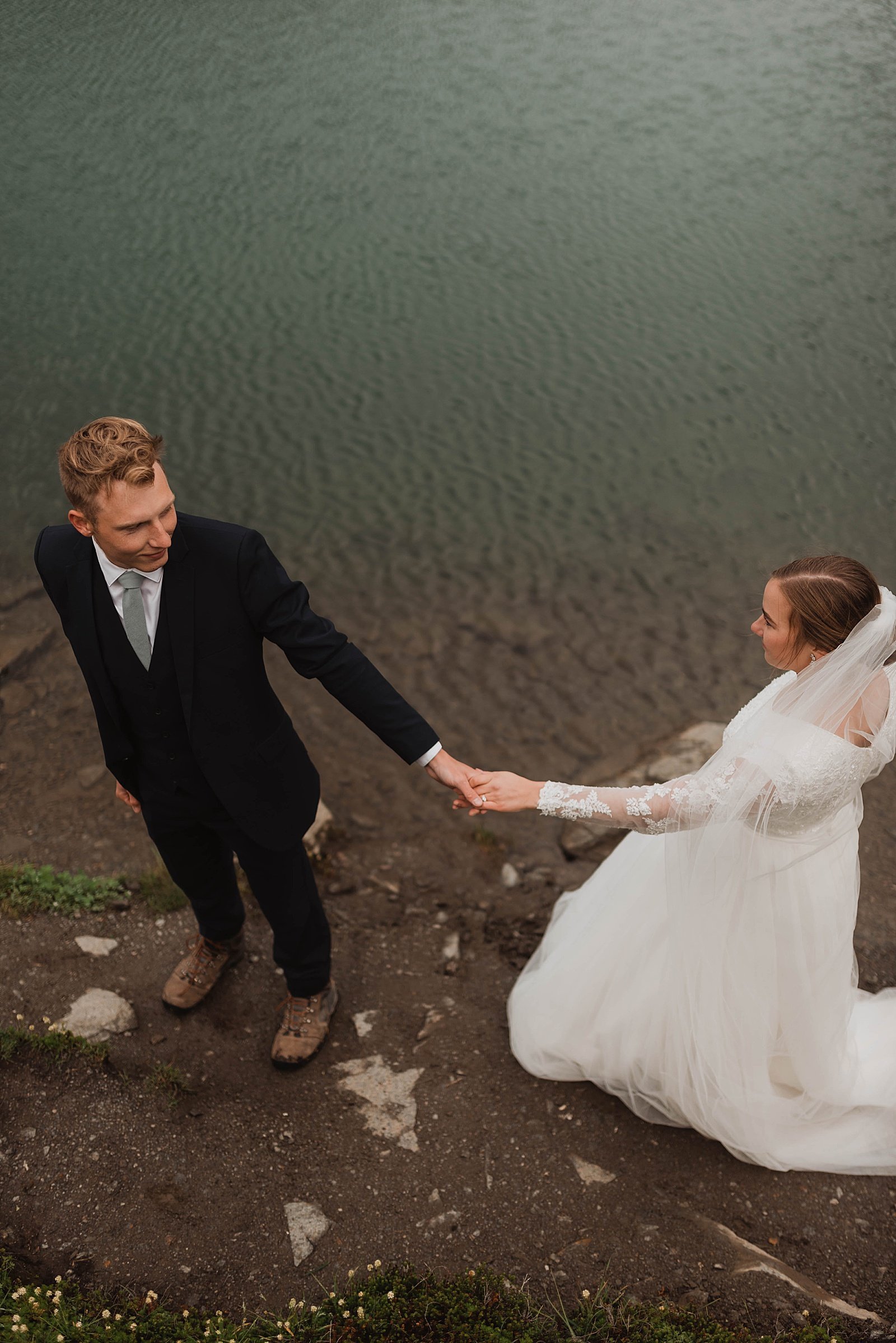  Newlyweds holding hands in front of a blue mountain lake at Hatcher Pass 