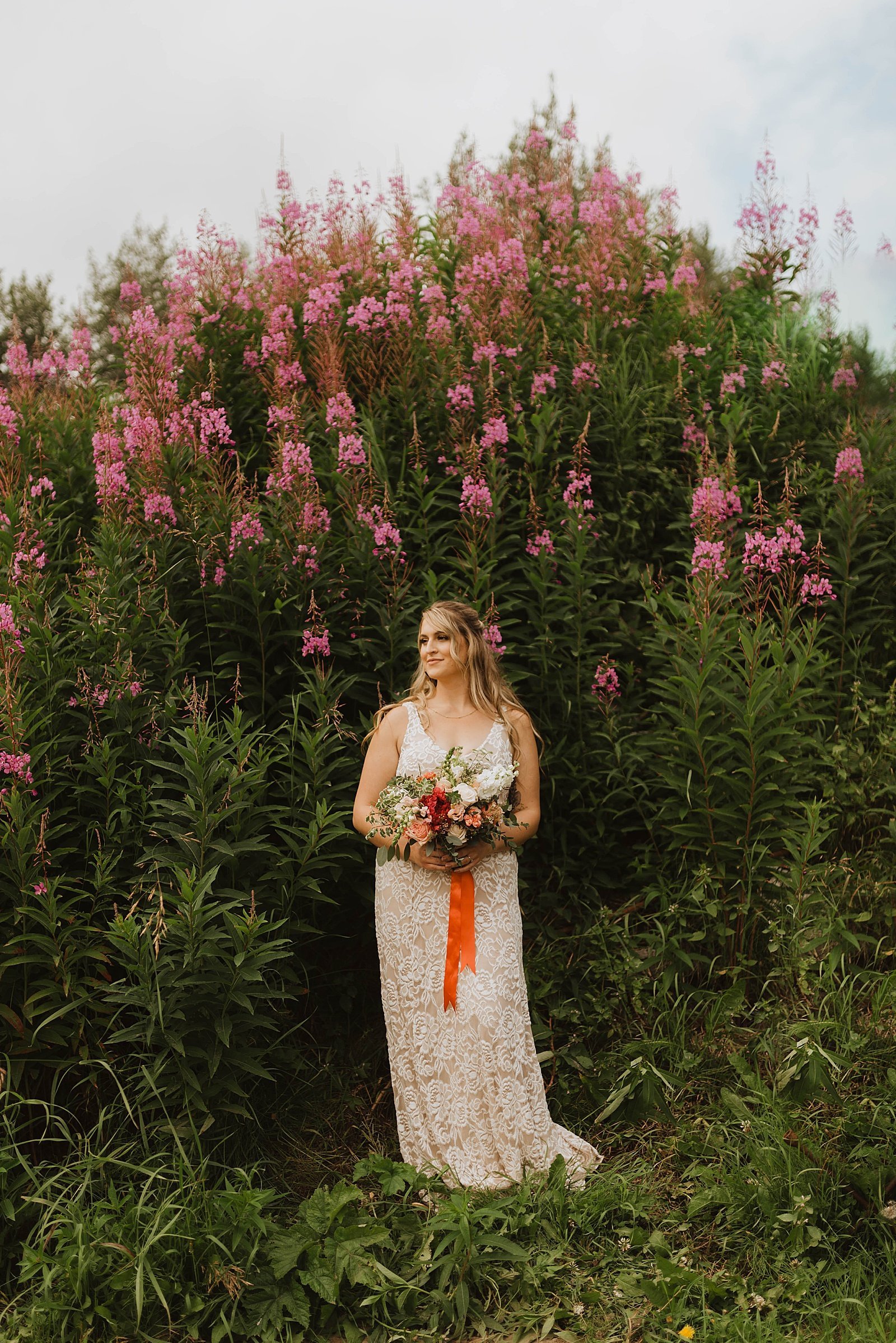  Boho bride in lace dress standing in front of tall red flowers for Alaska summer wedding.  