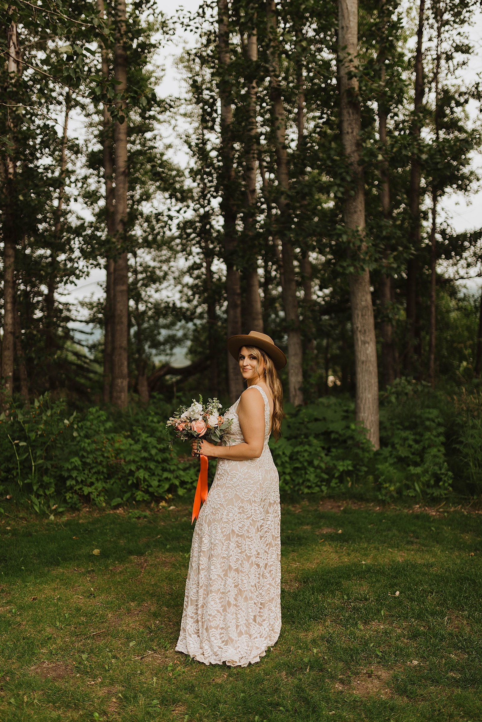  Alaska boho Summer bride in front of large trees wearing a hat and holding large bridal bouquet. 