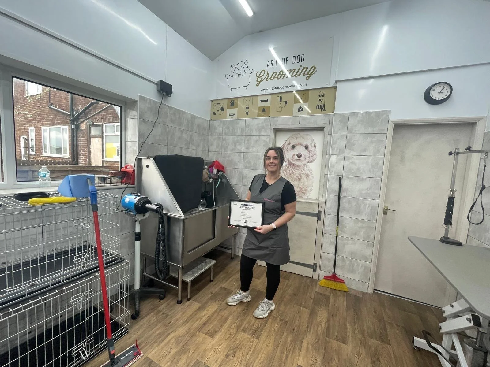 Woman in a dog grooming salon holding a certificate, with grooming equipment and kennels visible.