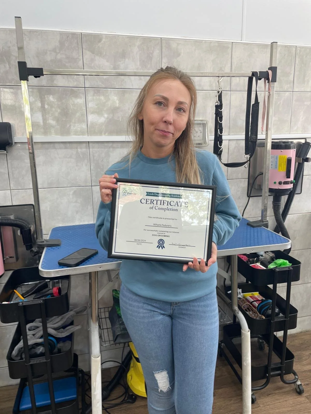 Woman holding a framed certificate of completion for dog grooming, standing by grooming equipment and tools in a tiled room.