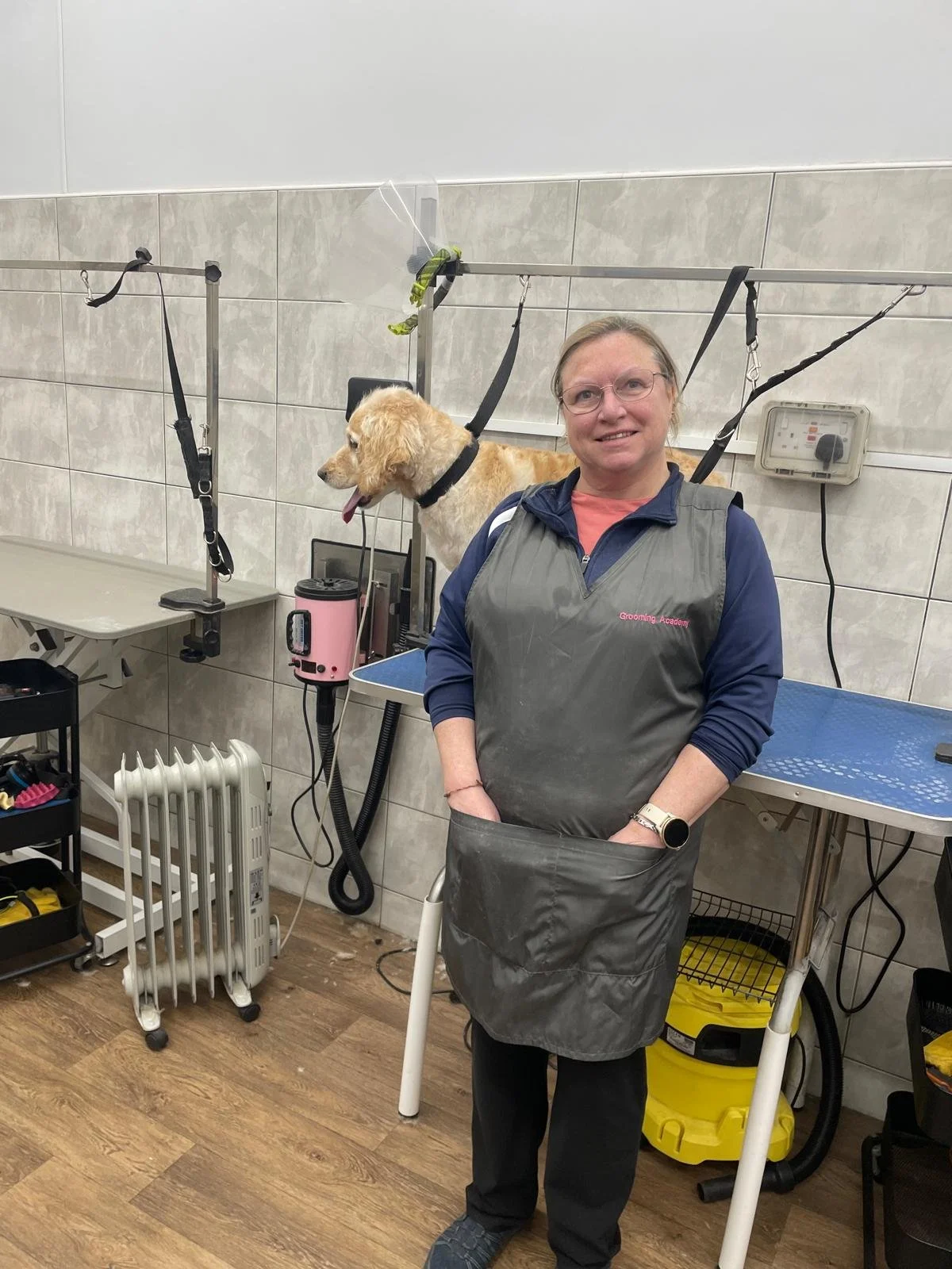 A woman standing in a pet grooming salon wearing a grooming apron, with grooming equipment around her and a dog on a grooming table in the background.