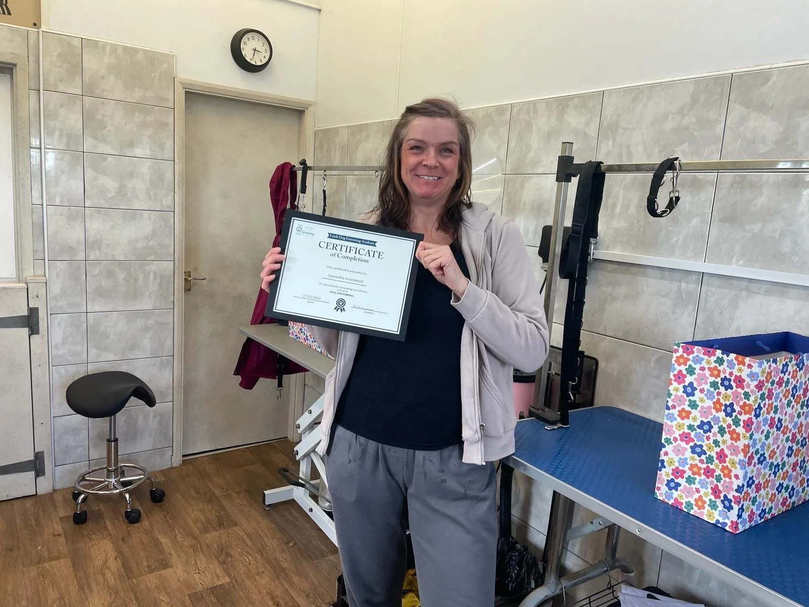 A person standing indoors holding a certificate with a smile. The room has a tiled wall, an adjustable grooming table, and a colorful gift bag.