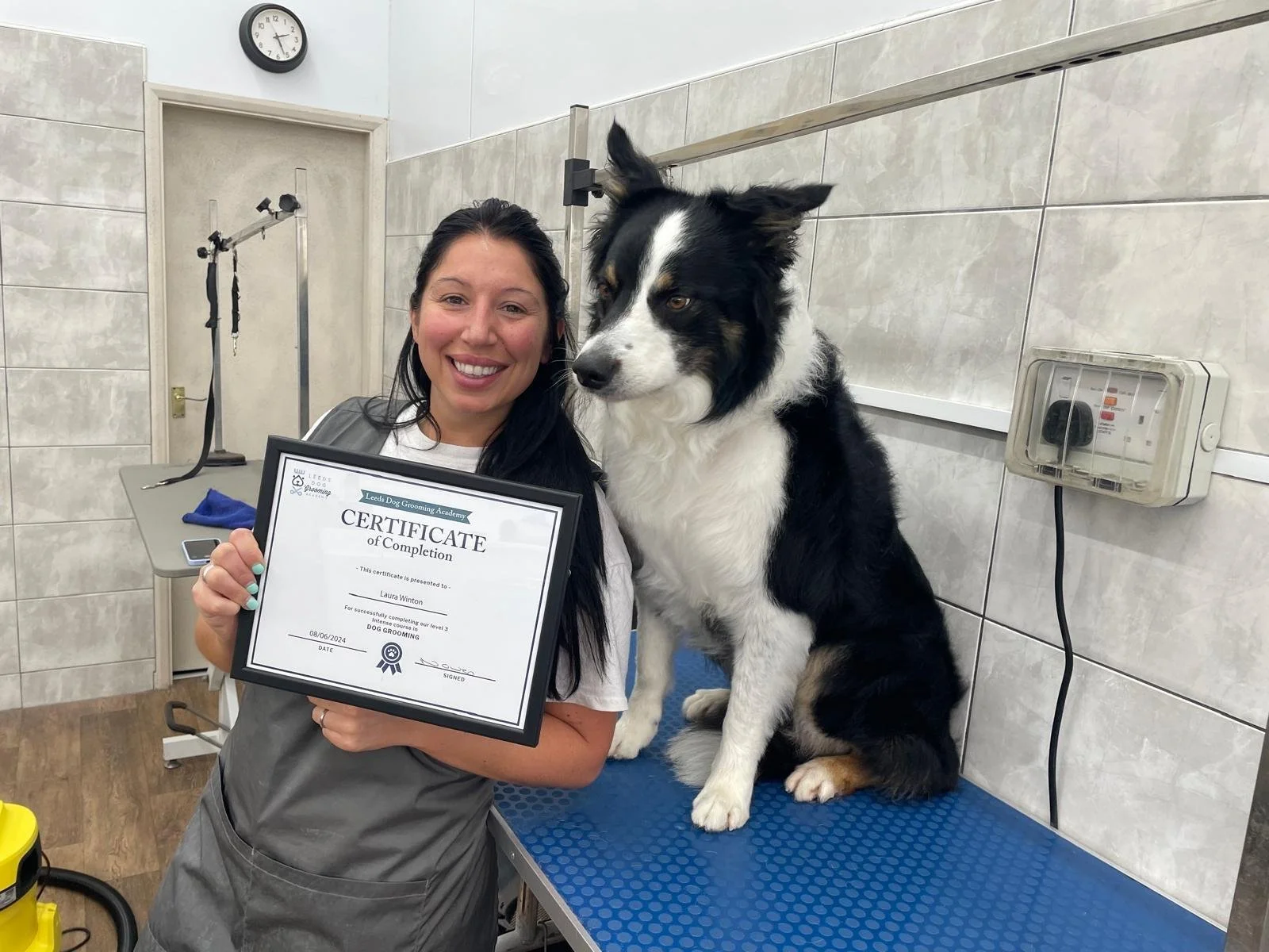 Woman holding a certificate next to a black and white dog on a grooming table in a pet grooming salon.