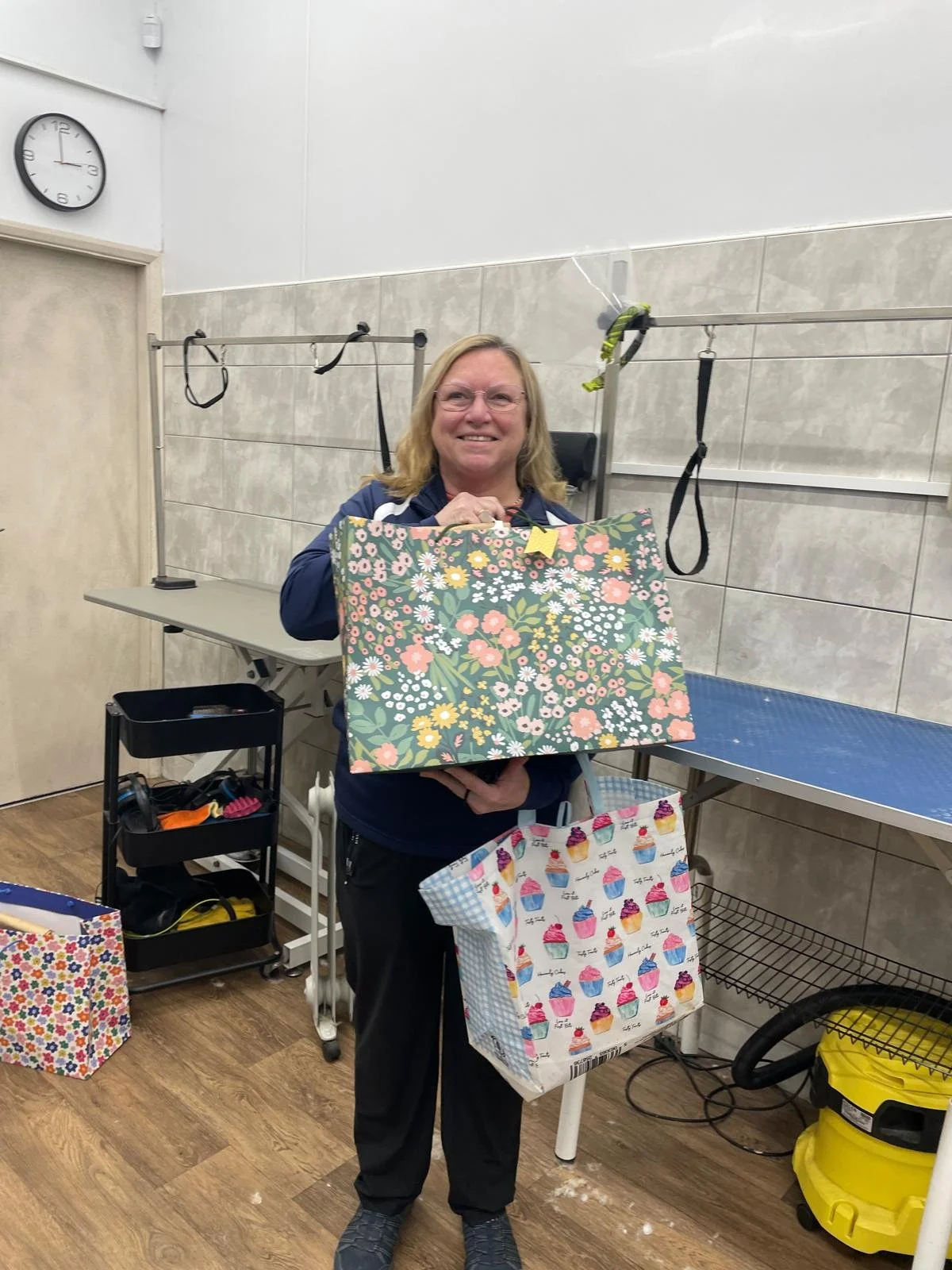 Person holding colorful gift bags in a room with grooming tables, a clock on the wall, and various equipment.