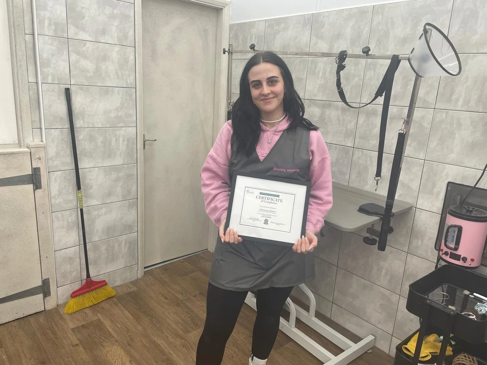 Woman holding certificate in grooming academy uniform in a grooming salon with a broom and grooming equipment nearby.