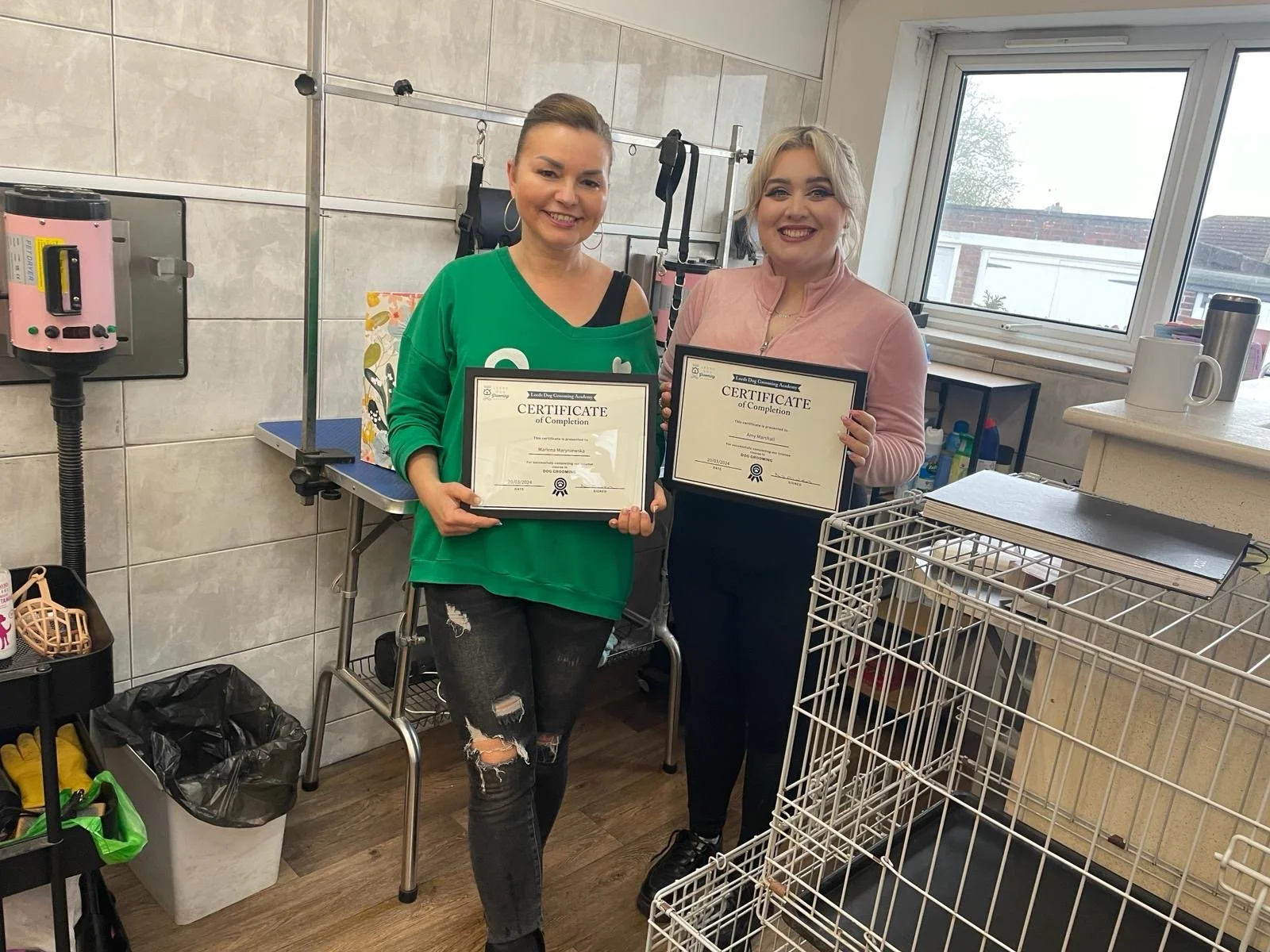 Two women holding certificates of completion in a grooming salon setting, smiling, surrounded by grooming equipment and a dog crate.