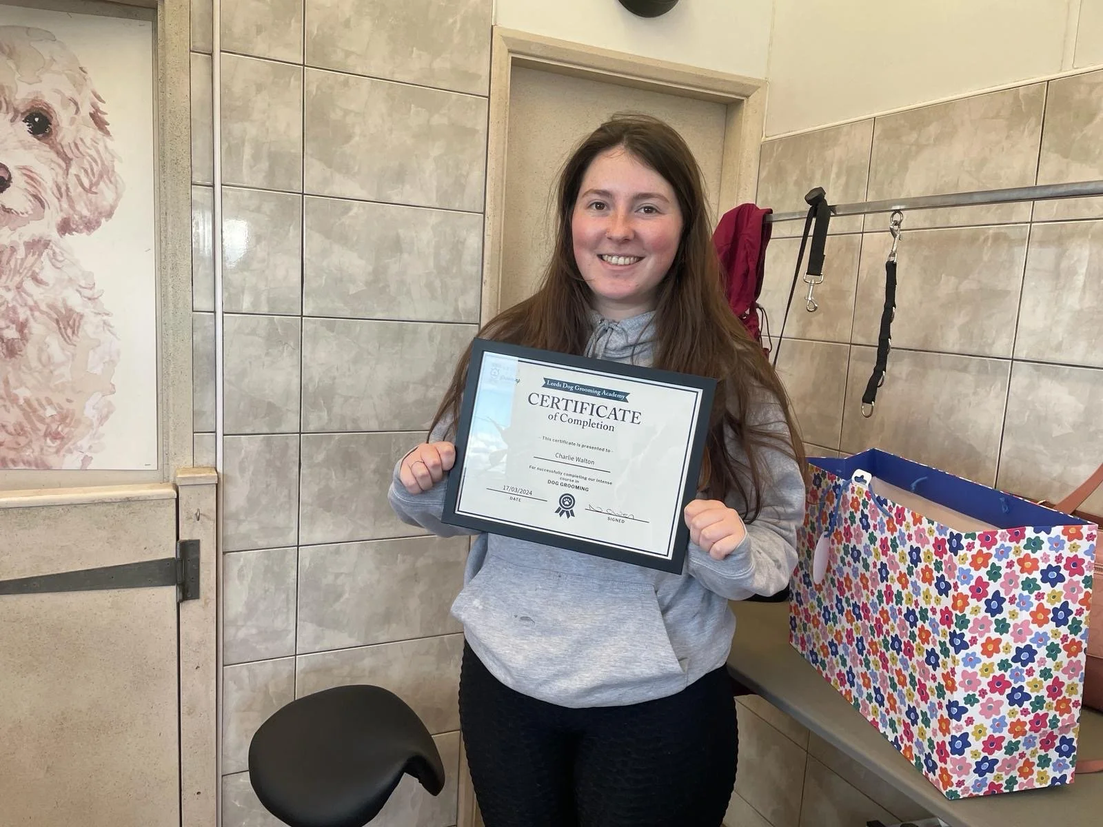 Woman smiling while holding a Certificate of Completion, standing in a room with tiled walls and a floral gift bag on a table.