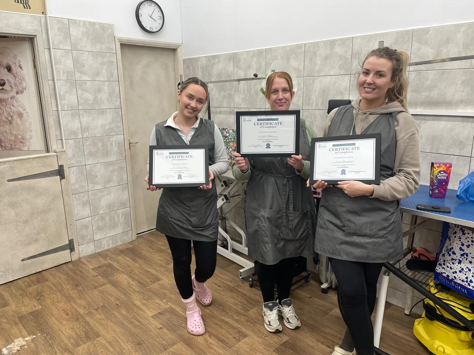 Three women wearing aprons stand in a room holding certificates. They are smiling, and the room features a wall clock and a partially visible image of a dog. The floor is wooden, and there's a table with various items, including a colorful bag and a
