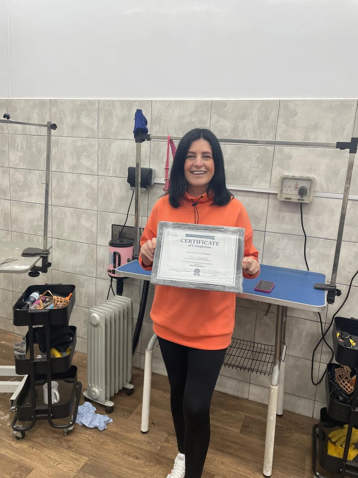 A person in an orange hoodie holding a certificate of completion in a grooming salon with equipment and tools visible in the background.