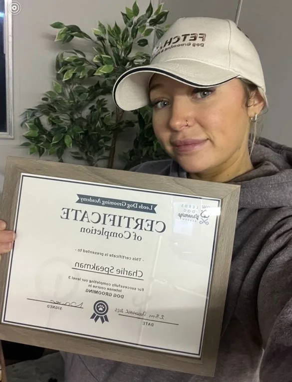 Person holding a certificate of completion from a dog grooming academy, wearing a cap and hoodie, standing in front of a plant.
