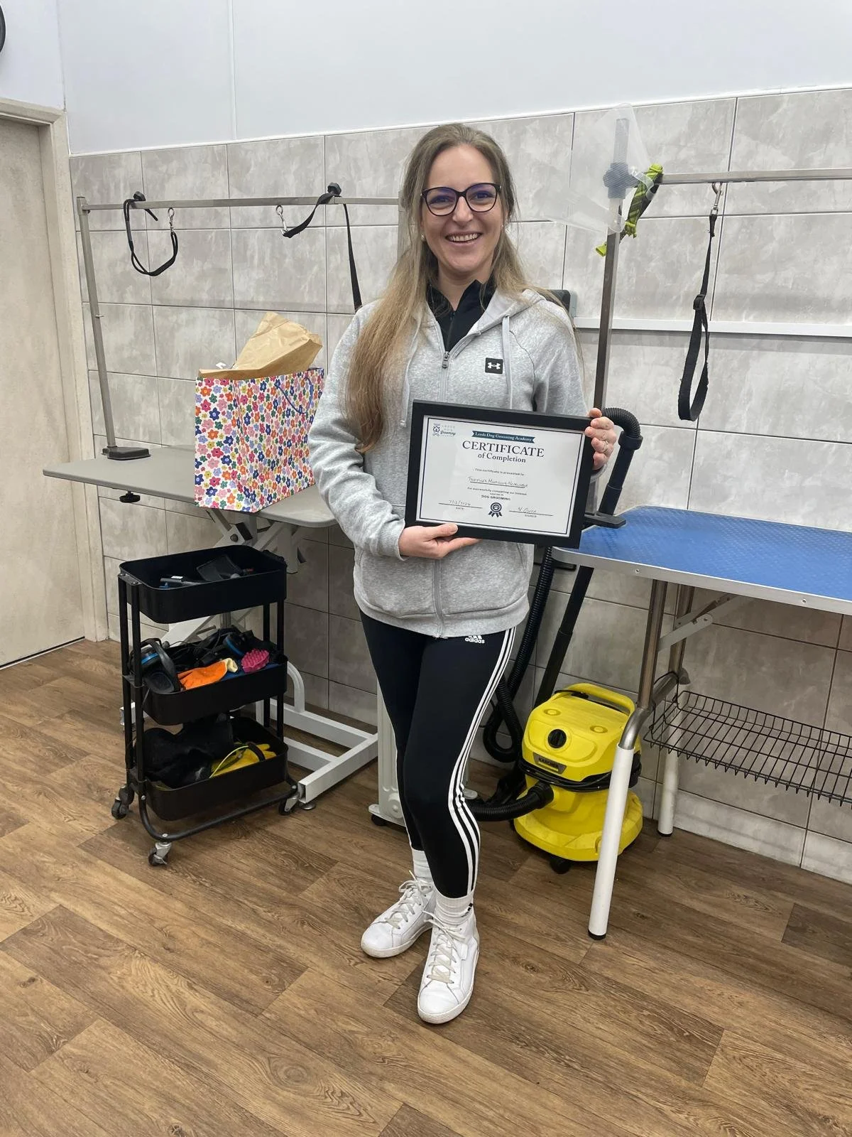 Person holding a certificate in a grooming salon, standing next to a grooming table with a colorful gift bag and grooming tools in the background.