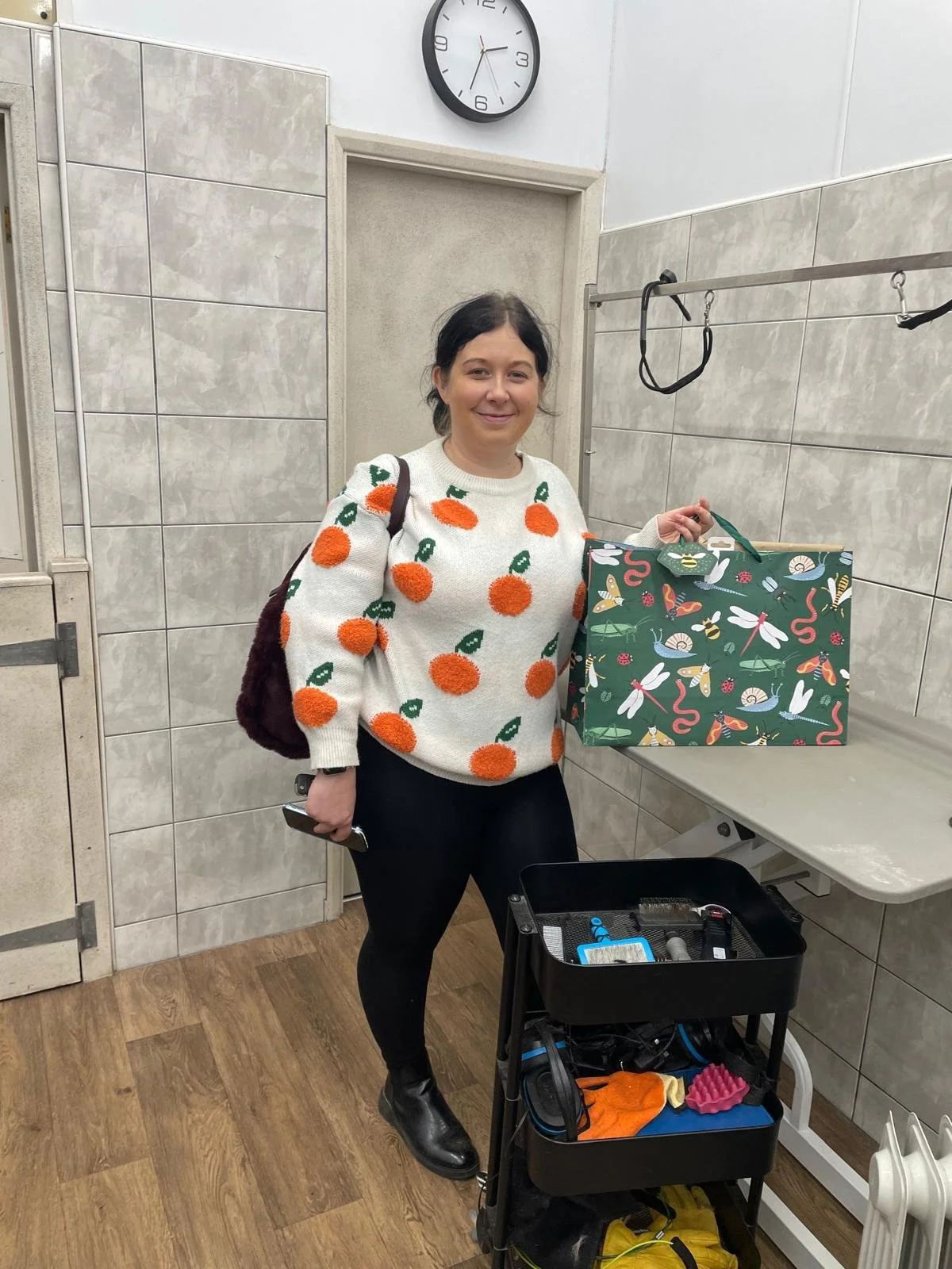 Woman in a room with tiled walls, wearing a sweater with orange fruit pattern, holding a decorative bag, standing beside a cart with supplies, under a wall clock.