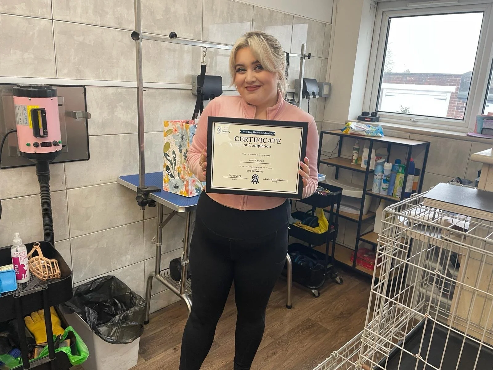 Person holding a certificate in a pet grooming salon, standing next to grooming equipment and supplies.