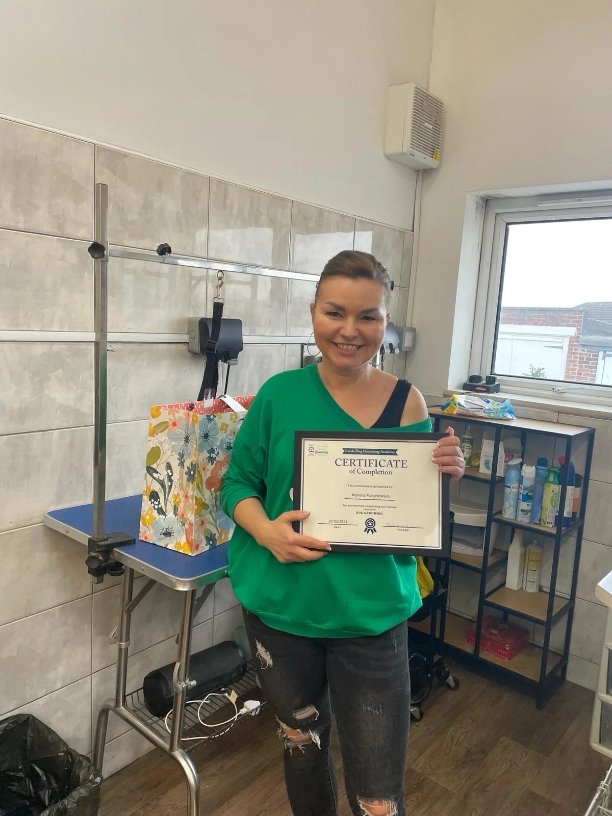 Woman holding a certificate of completion in a room with grooming supplies, standing next to a colorful gift bag.