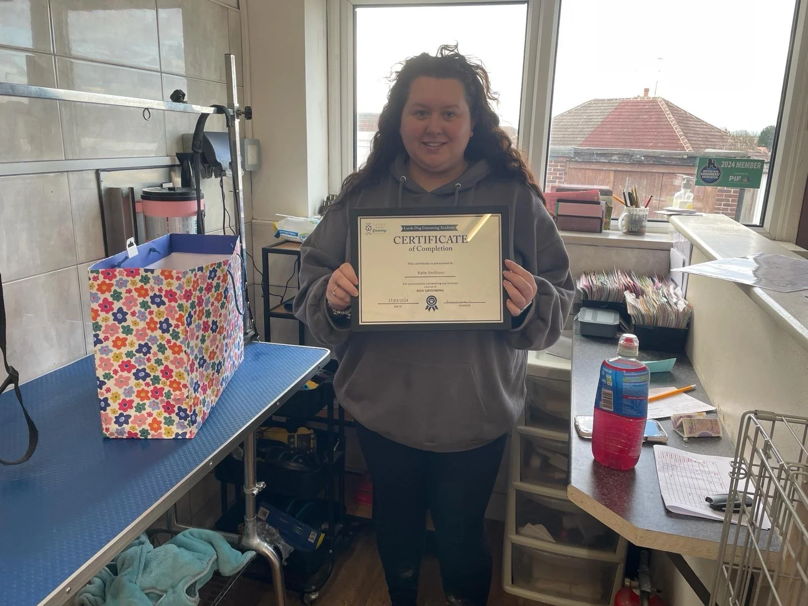 Person holding a certificate in a grooming salon with a floral gift bag on a table.