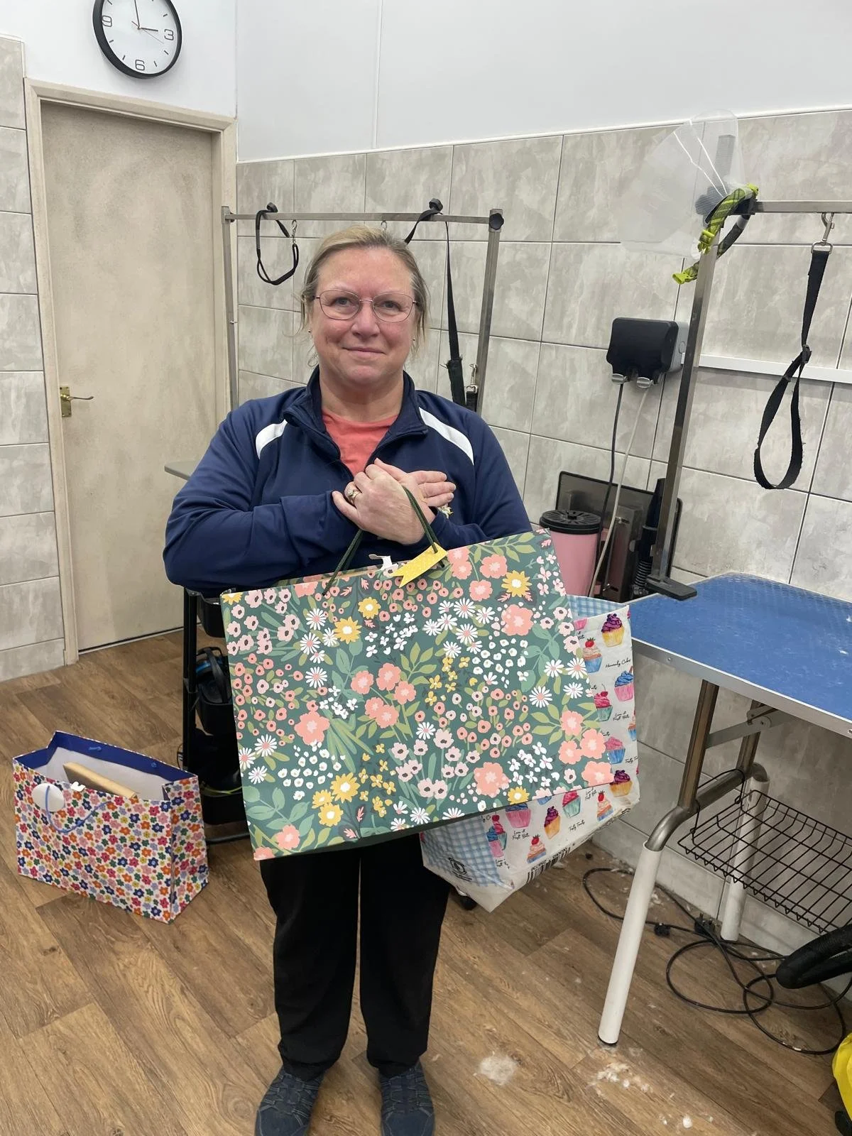 Woman in a room holding a floral gift bag, smiling, with grooming equipment in the background.