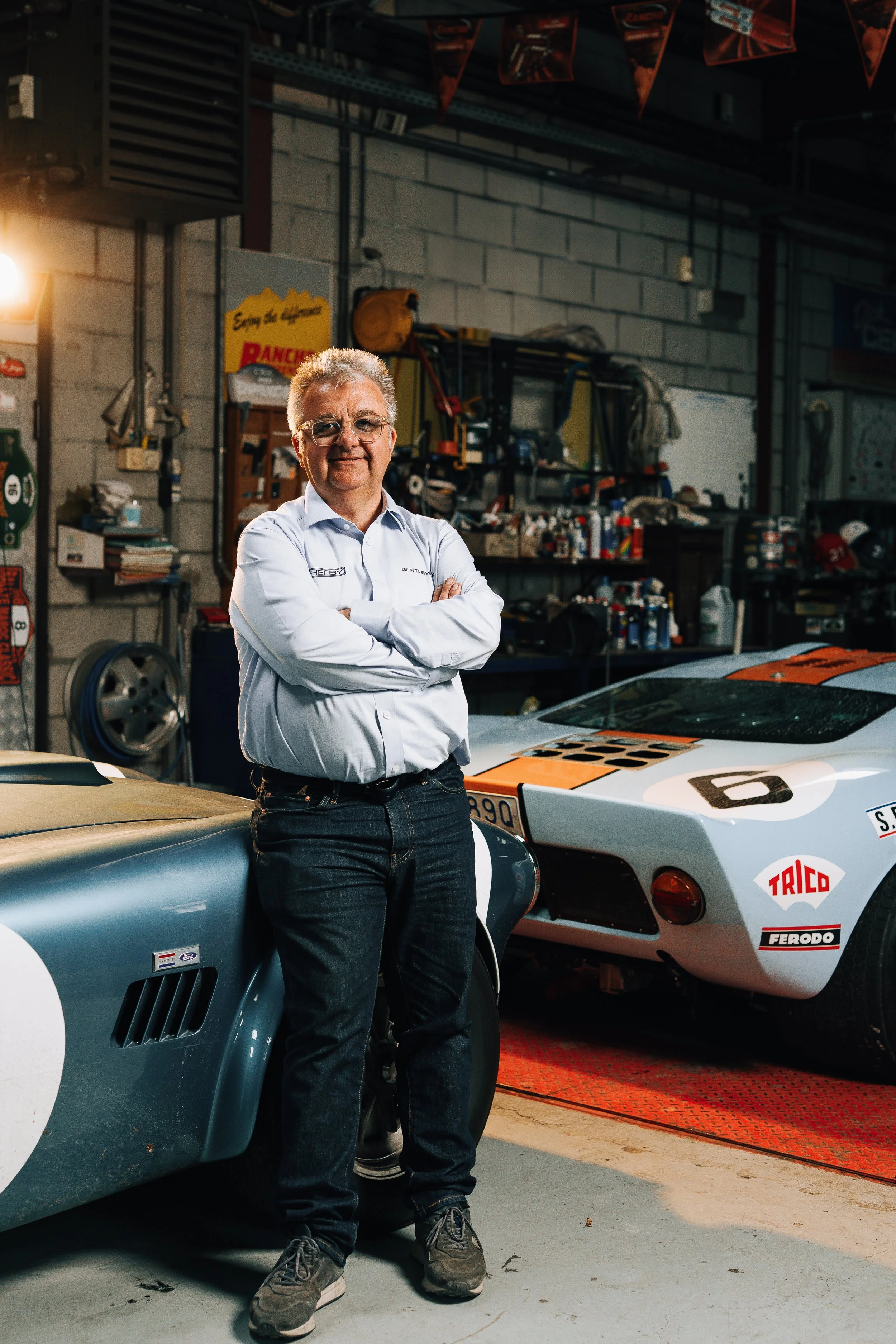 A man with gray hair and glasses in a white shirt stands with arms crossed in a garage, surrounded by vintage race cars and various tools.