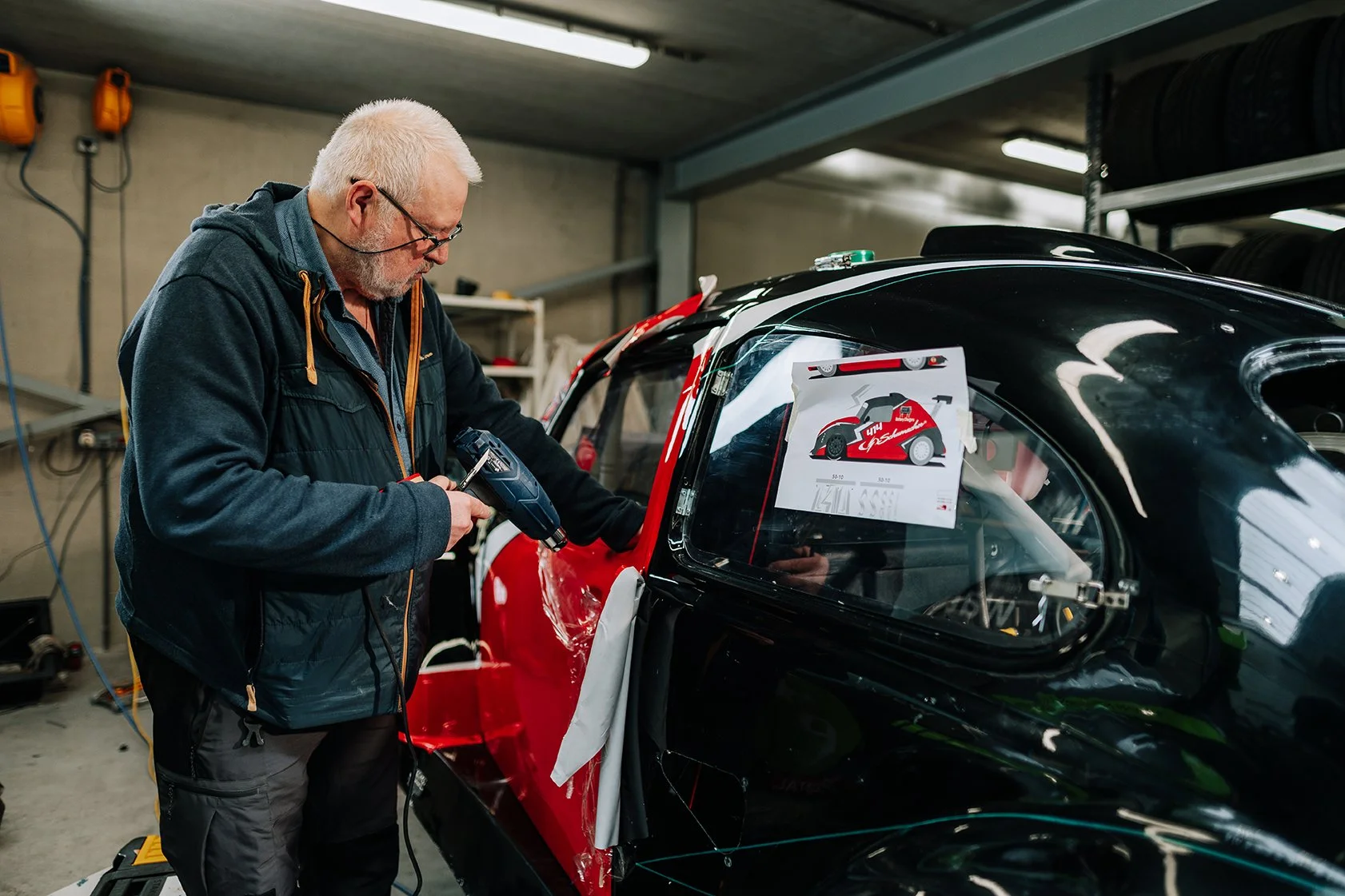 A man working on a race car in a garage, using a power tool, with racing posters on the window.