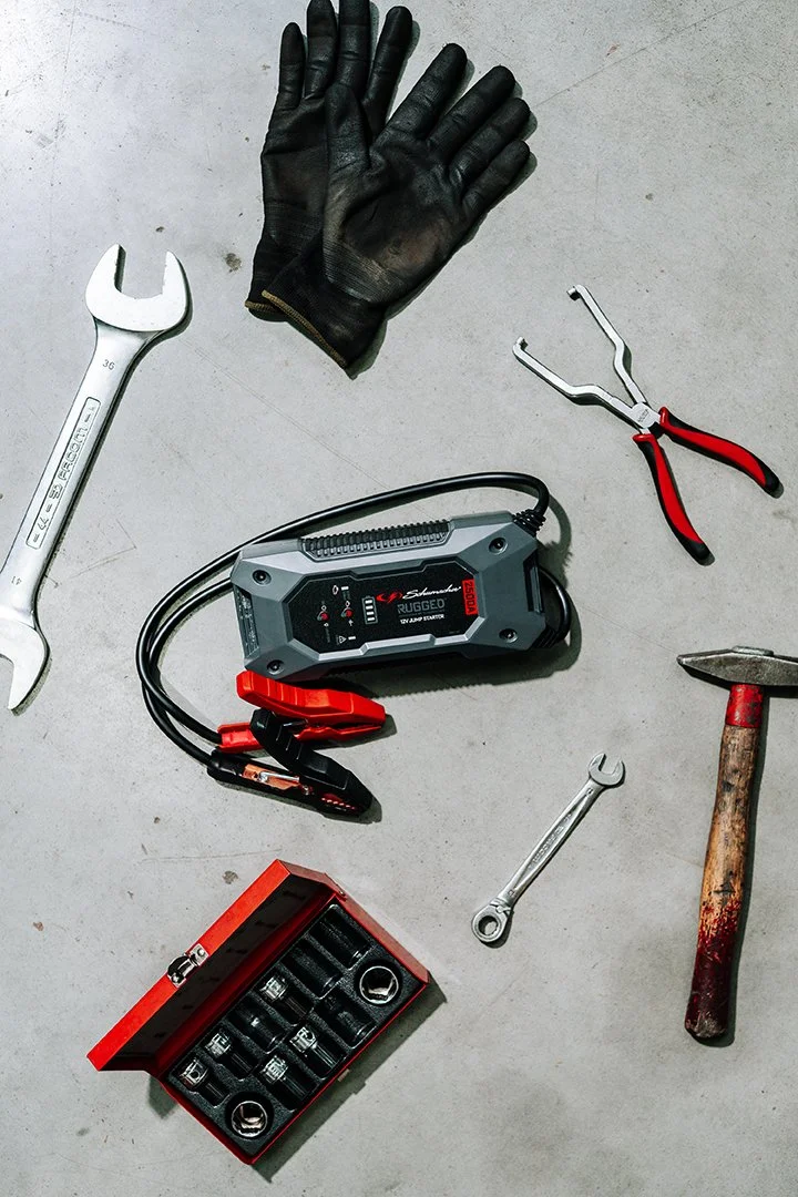 A set of tools and equipment on a concrete surface, including a pair of black work gloves, a wrench, a pair of pliers, a digital battery tester, a small socket set, a wrench, and a hammer with a wooden handle.