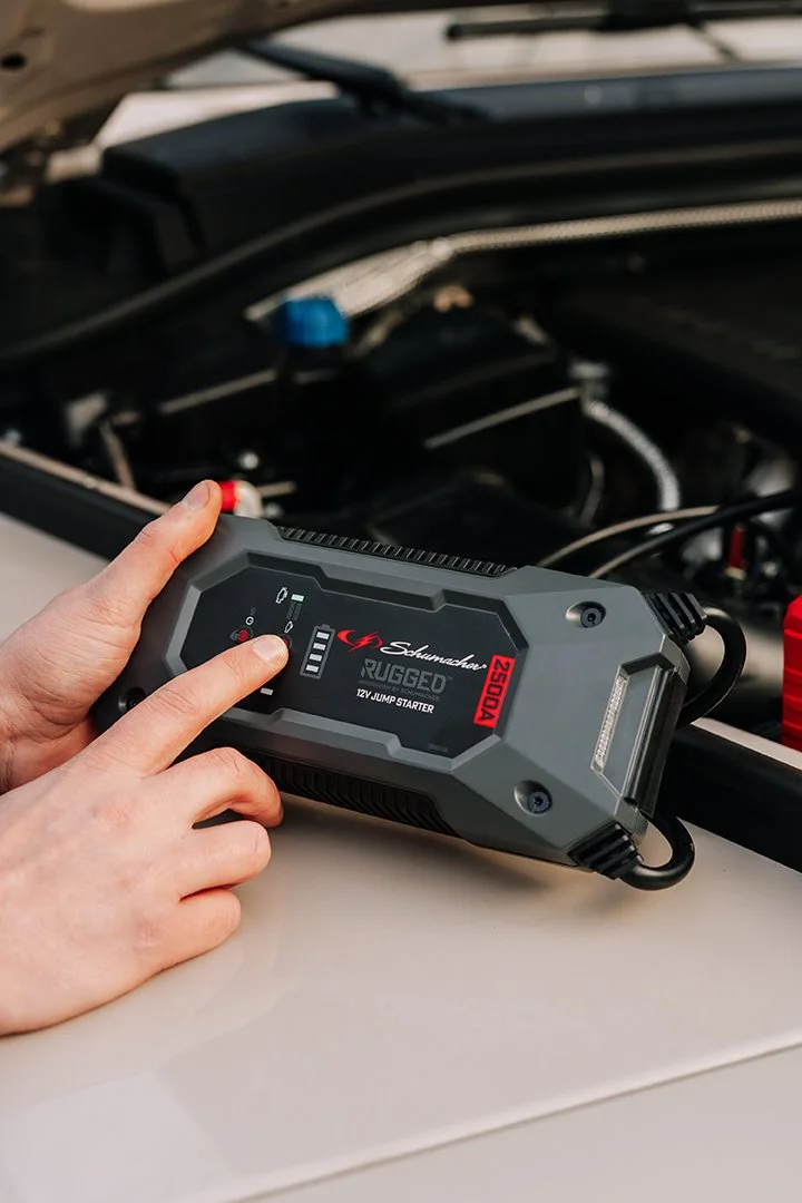 Person using a Schumacher Rugged 12V jump starter to boost a car with the hood open.