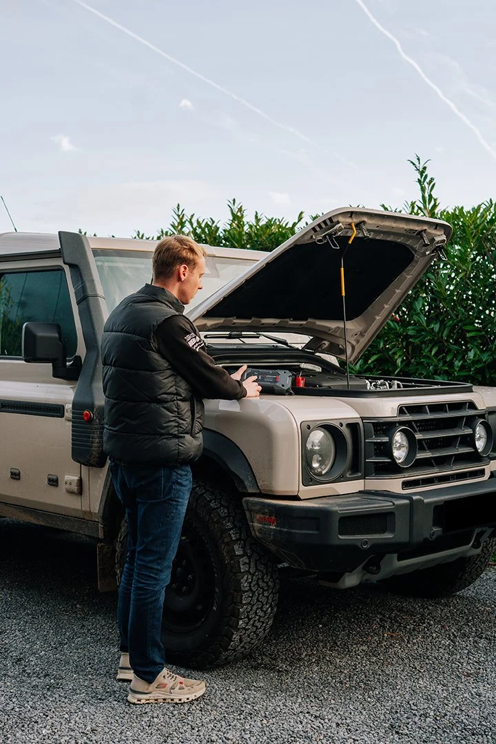A man checking under the hood of a beige off-road vehicle parked outdoors on a gravel surface.