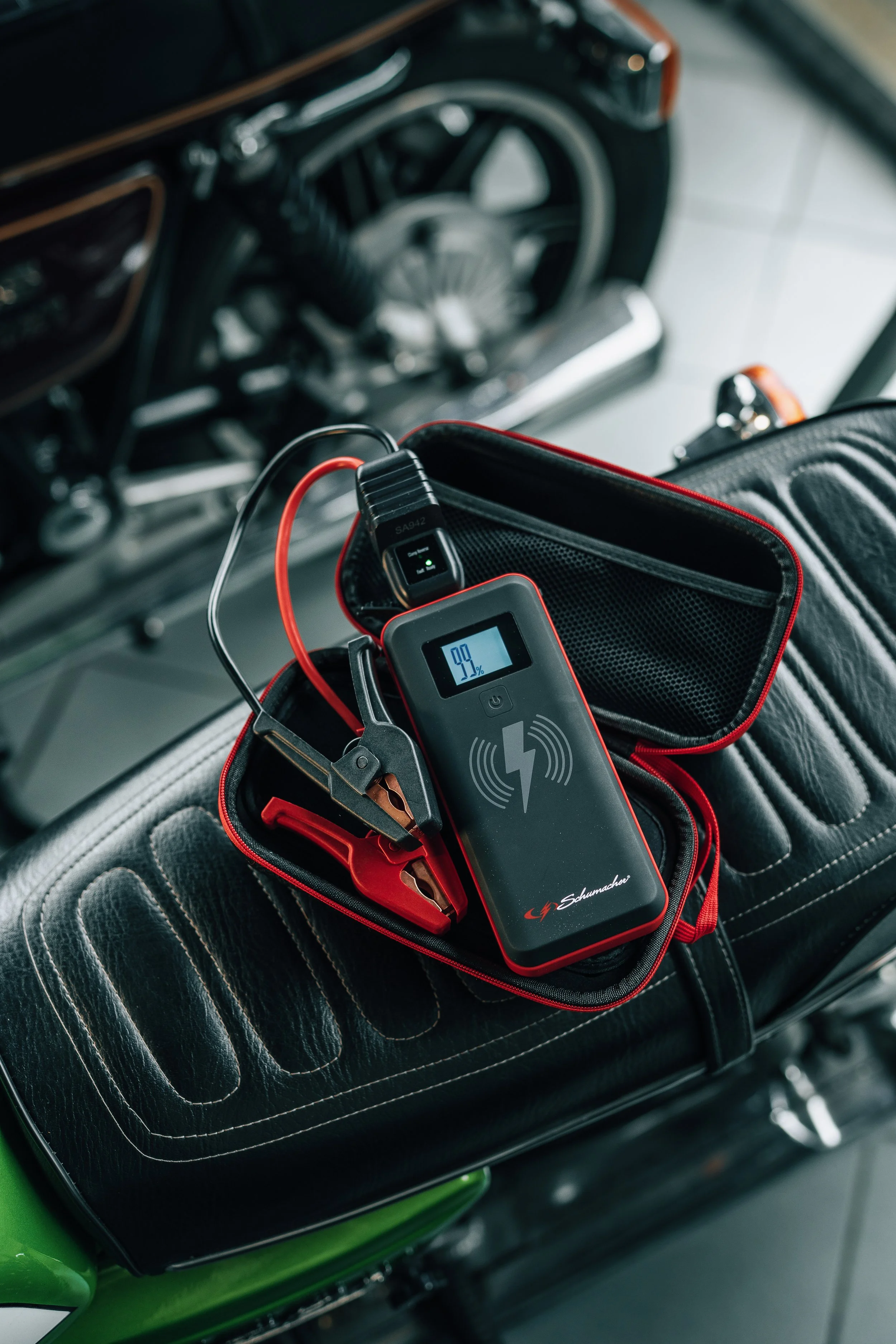 A portable battery charger with a displayed 99% charge, connected to a vehicle, resting on a textured black seat with a red and black cover. The charger has a lightning bolt symbol and a wireless charging icon on its surface.