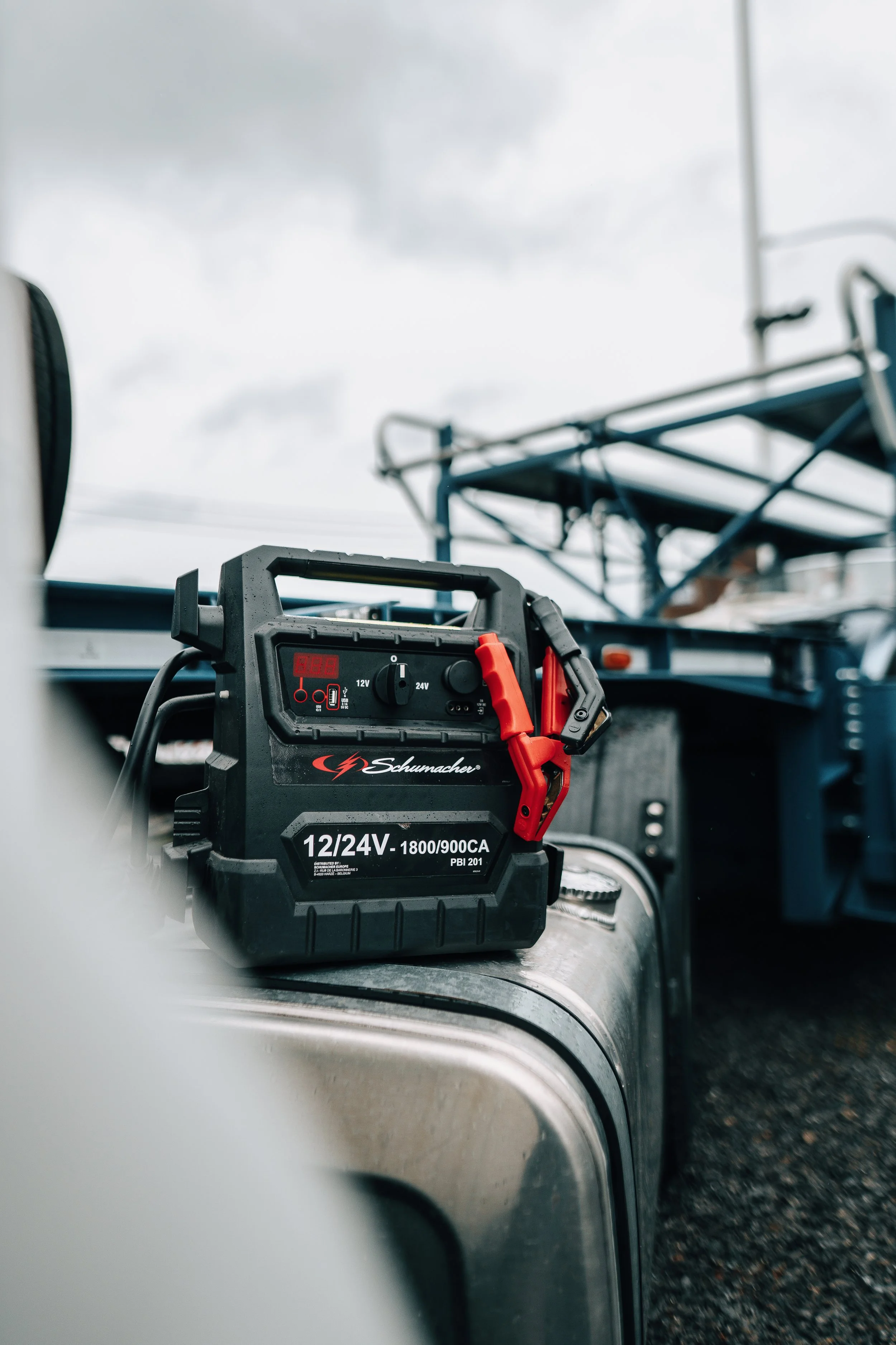 A digital battery charger placed on a metal surface, with a blurred background of a garage or workshop setting.