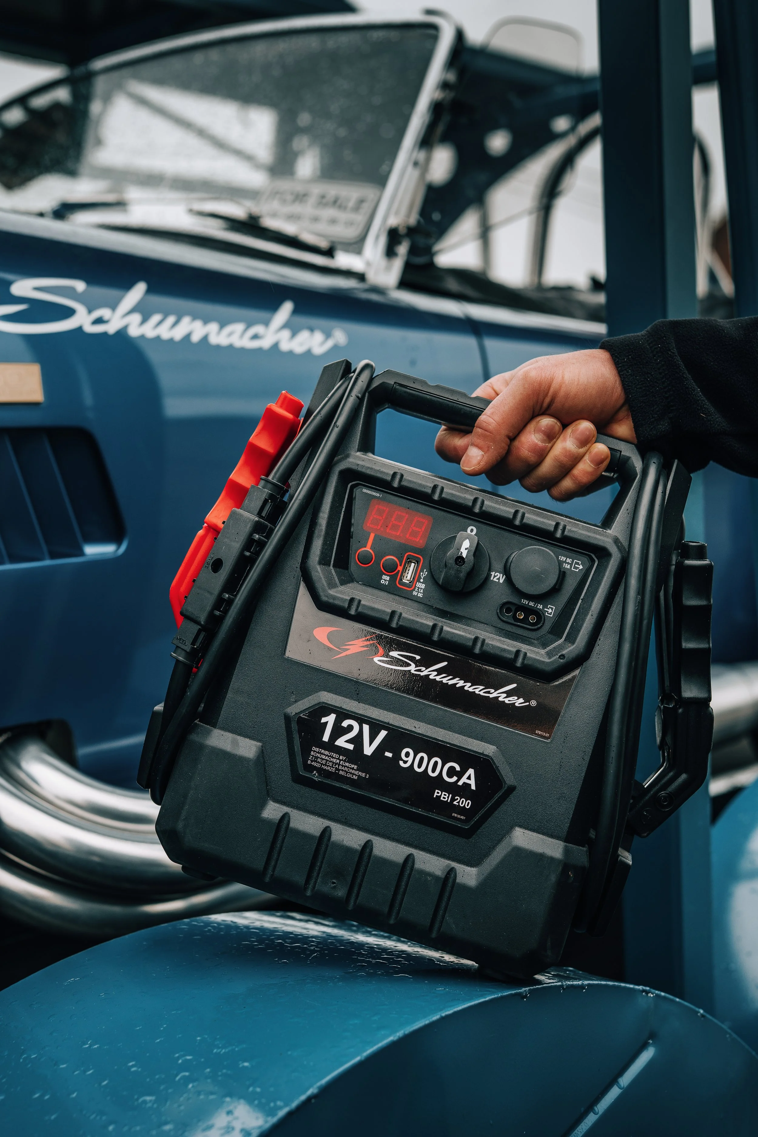 Close-up of a handheld jump starter with a digital display, red and black cables, and brand name 'Schumacher' in front of a blue motorcycle with the name 'Schumacher' on the tank.