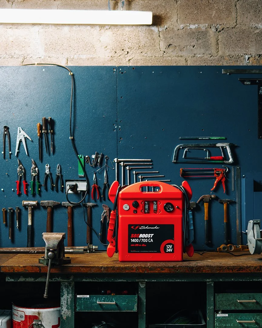 A workshop with a blue pegboard wall holding various hand tools, a large red battery jump starter, and a workbench with a vise, against a concrete wall.