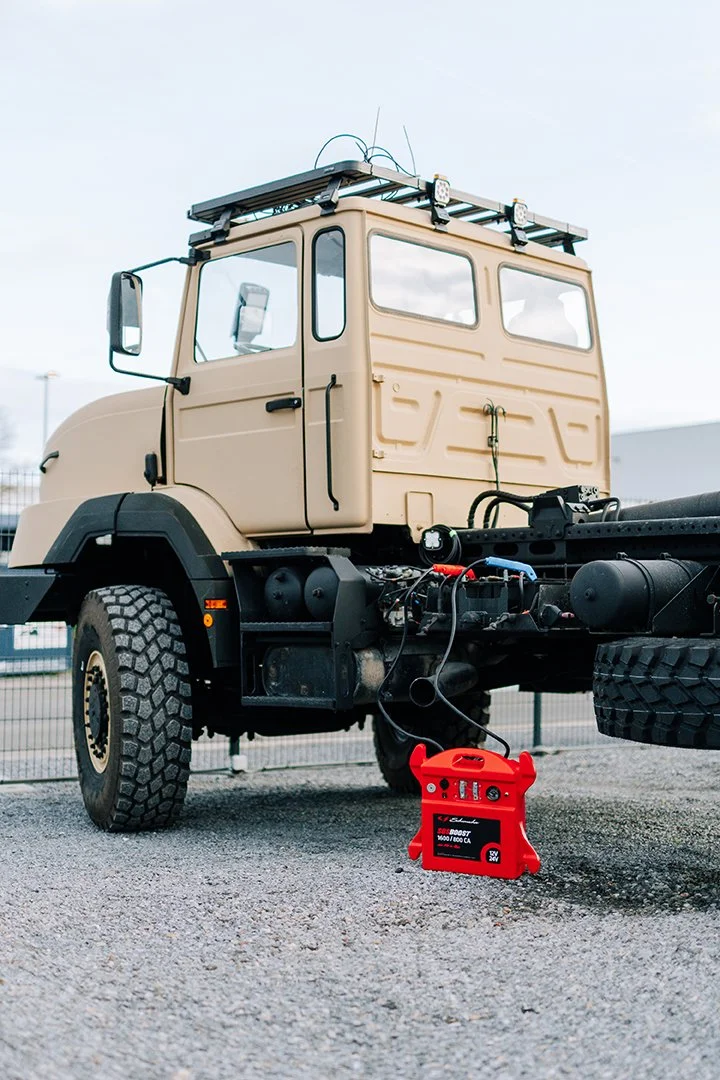 A beige truck parked outdoors with a ladder on top, connected to a red battery charger on the ground.
