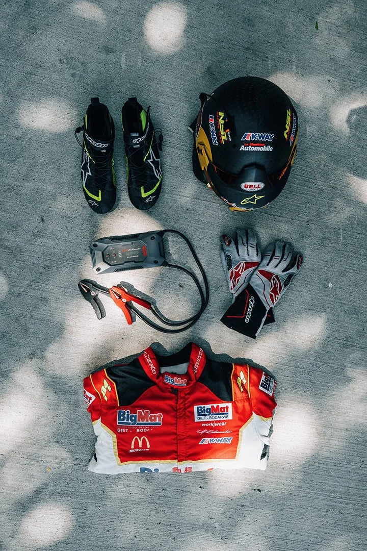 Racing helmet, racing gloves, racing shoes, car battery charger, racing suit with sponsor logos, all arranged on a concrete surface.