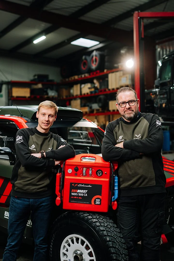 Two men standing in a garage next to a vehicle, holding a red portable jump starter. The garage has shelves filled with boxes and equipment, and the men are wearing matching black and green racing jackets.