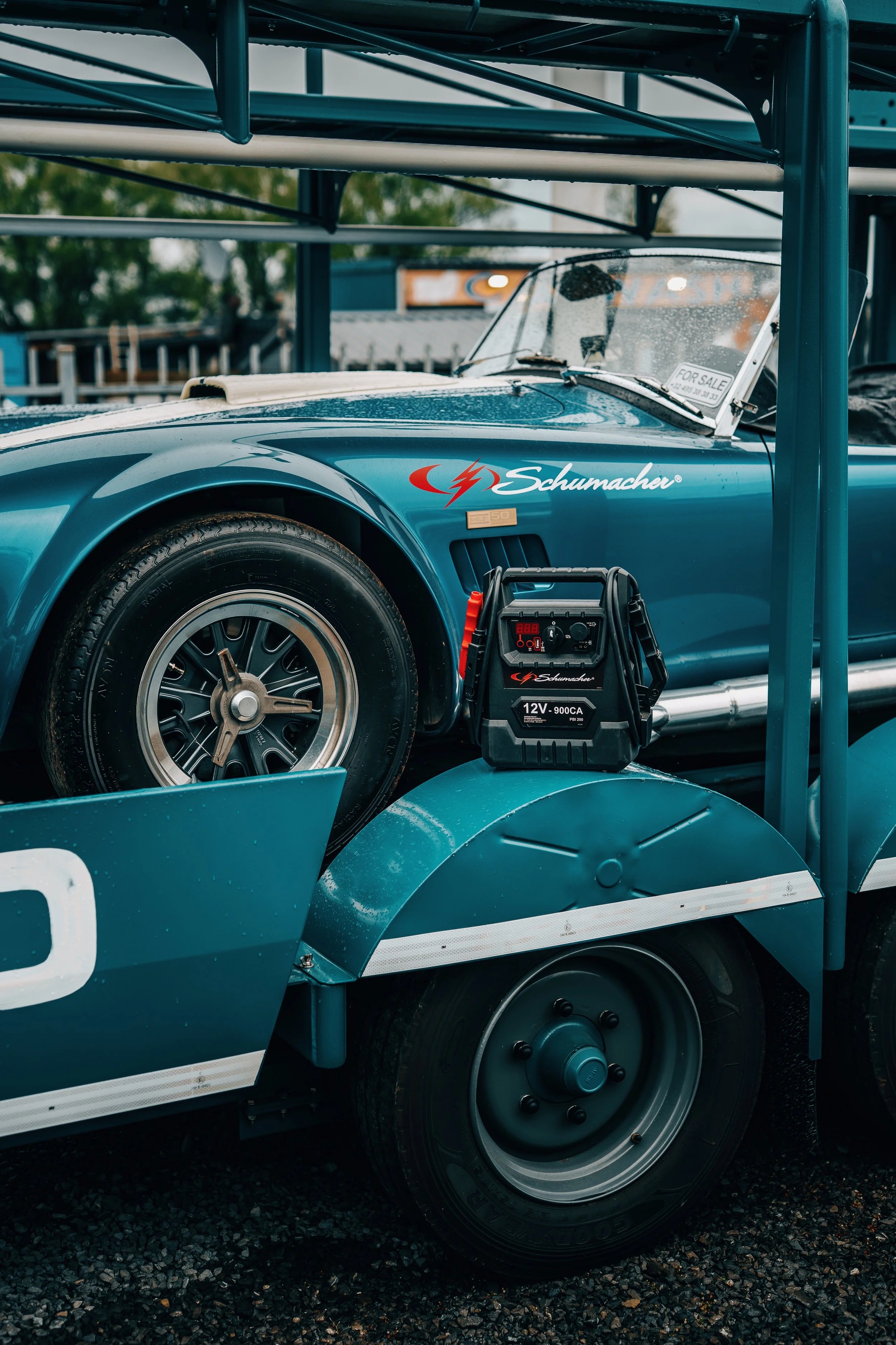 Close-up of a vintage sports car on a delivery truck, with a portable battery pack placed near the wheel, and a 'For Sale' sign in the windshield.