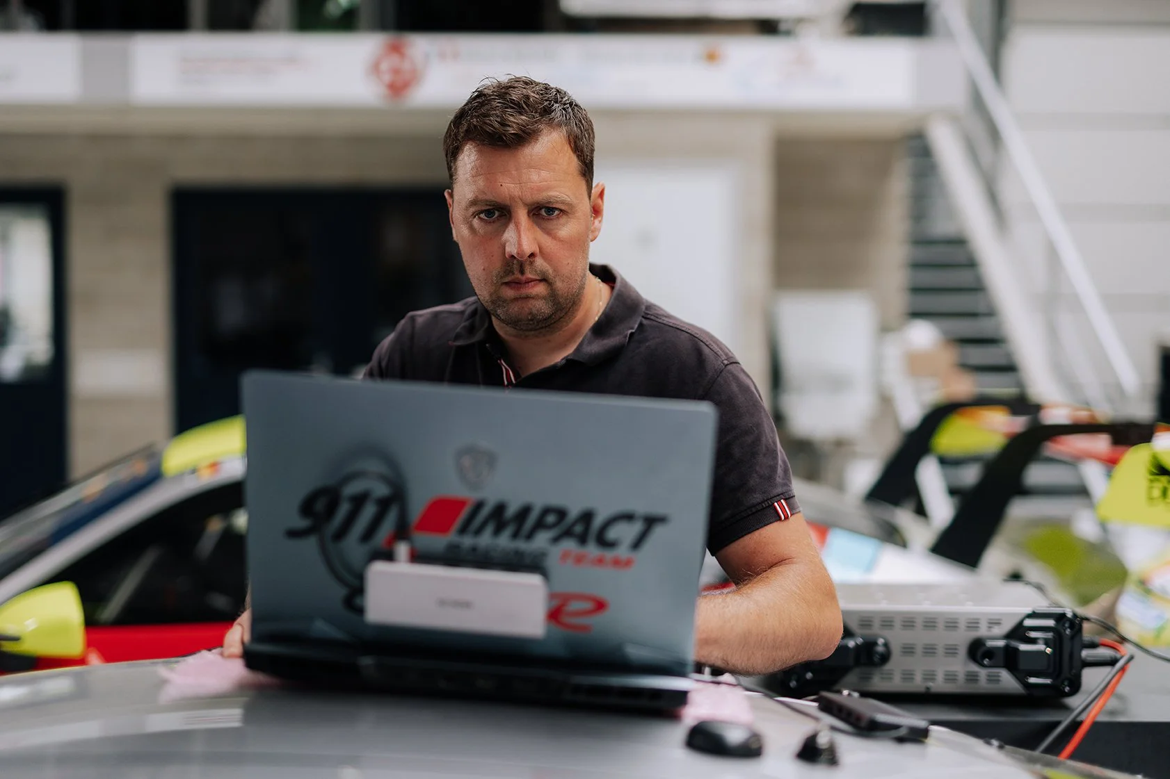 A man with short brown hair and a serious expression working on a laptop at a table covered with electronic equipment, with racing bikes and a building in the background.