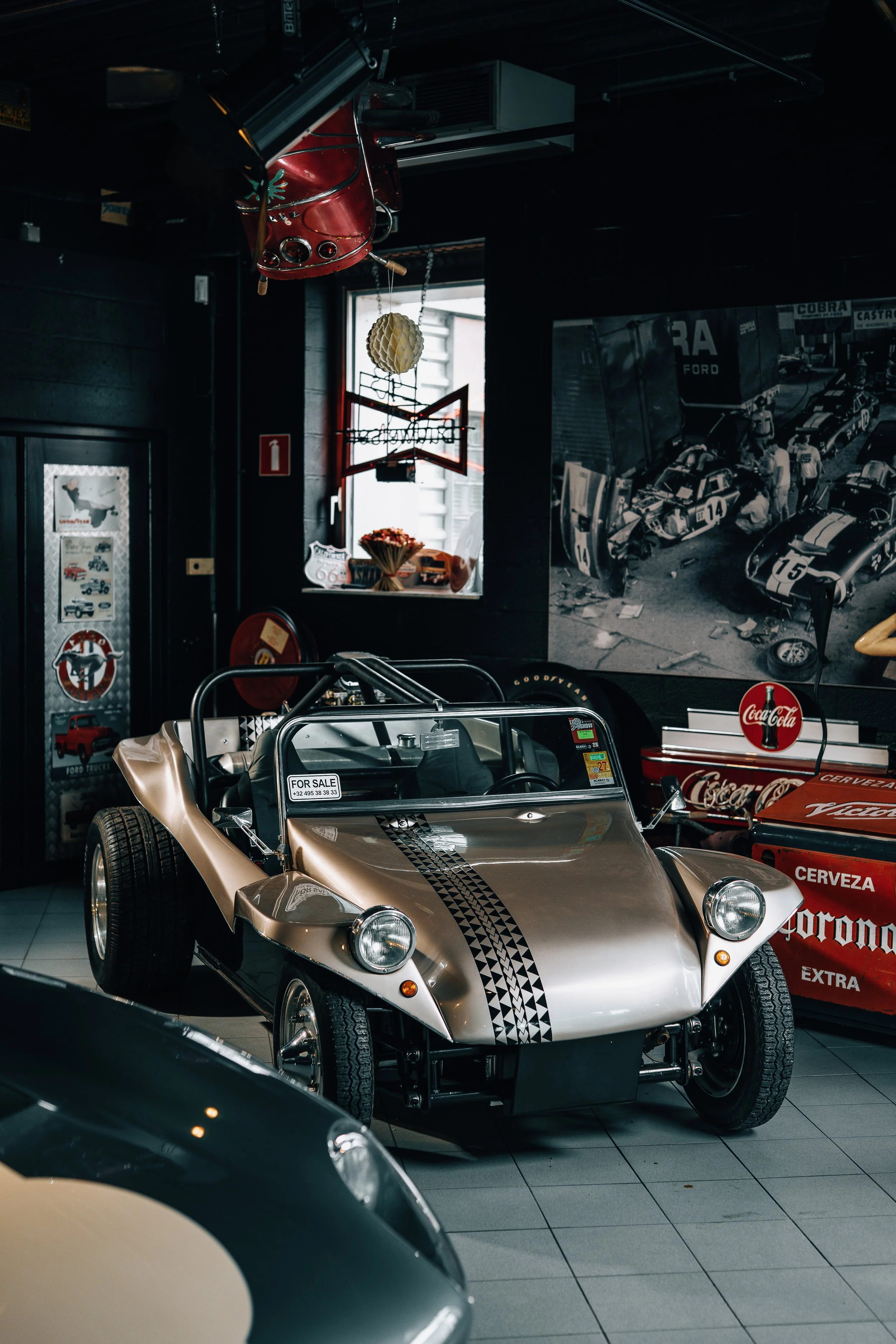 A vintage silver and black sports car with a checkered racing stripe parked indoors. Behind it, there are various racing-themed posters and memorabilia, including a large black-and-white photo of race cars and a display case with model cars. The room
