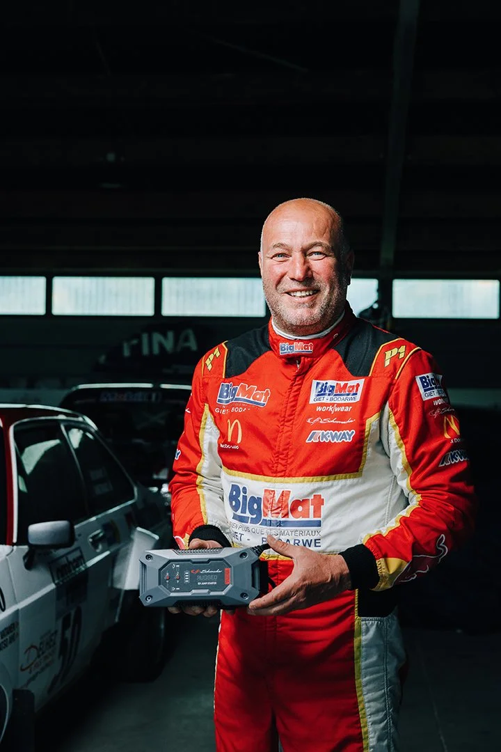 A smiling man in a red racing suit holding a portable jump starter in a garage with racing cars in the background.