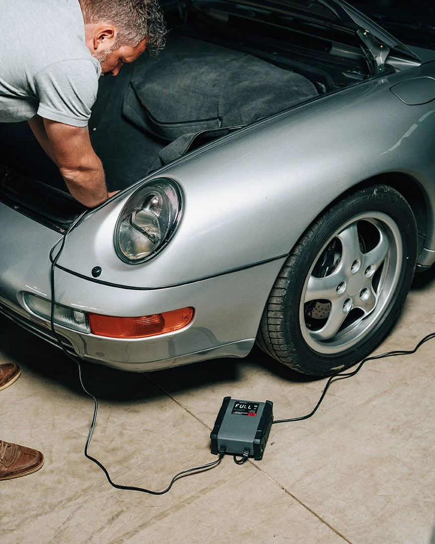 A man working under the hood of a silver Porsche sports car, connecting a diagnostic device to the vehicle.