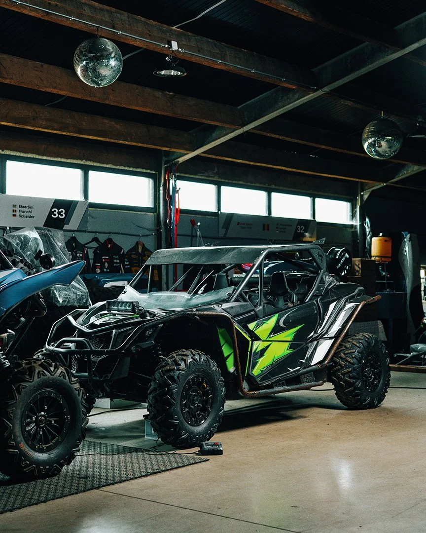 Off-road utility vehicle with green graphics, large rugged tires, and a roll cage inside a garage or showroom.