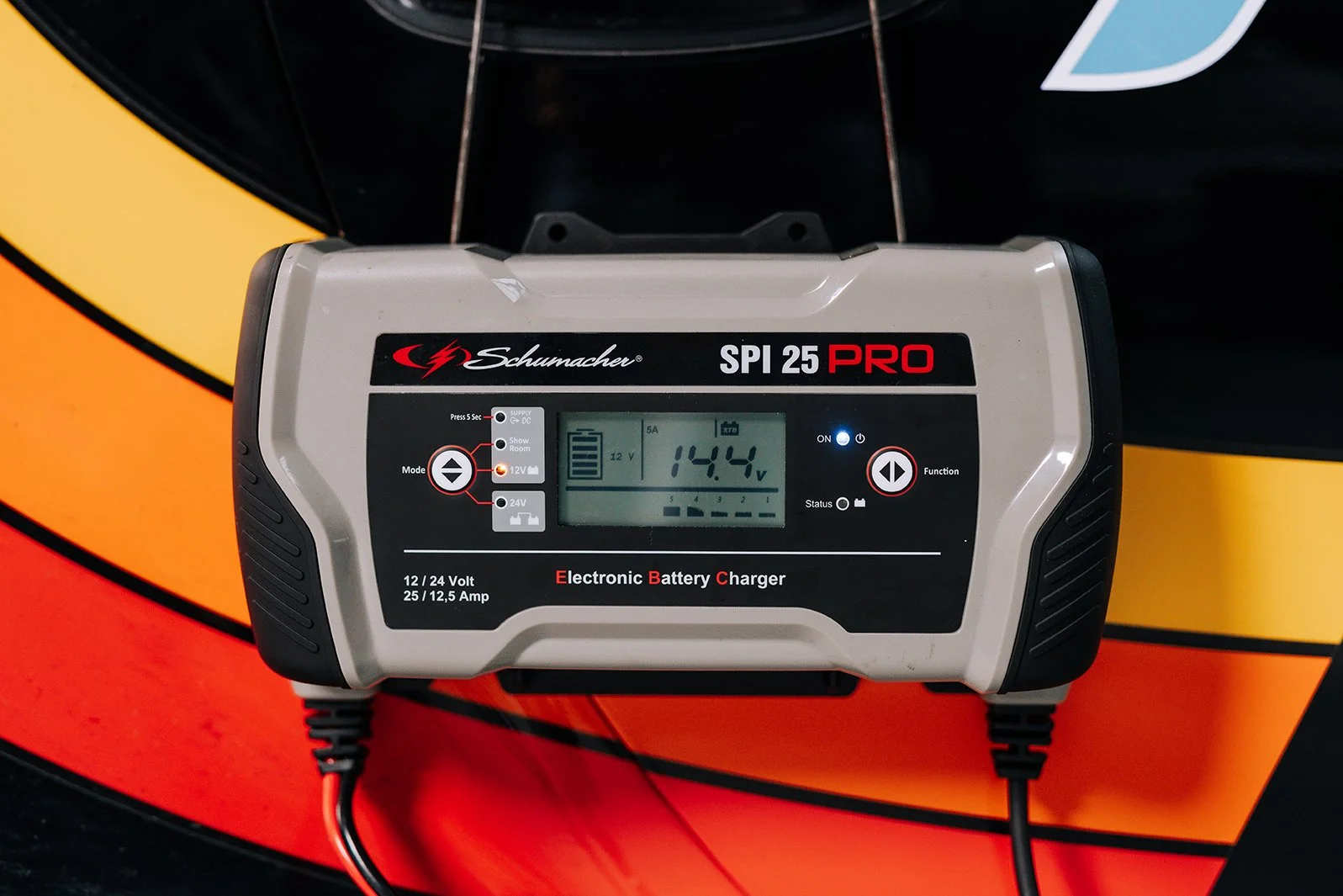 A Schumacher SPI 25 PRO electronic battery charger with a digital display showing 14.4 volts on a colorful background.