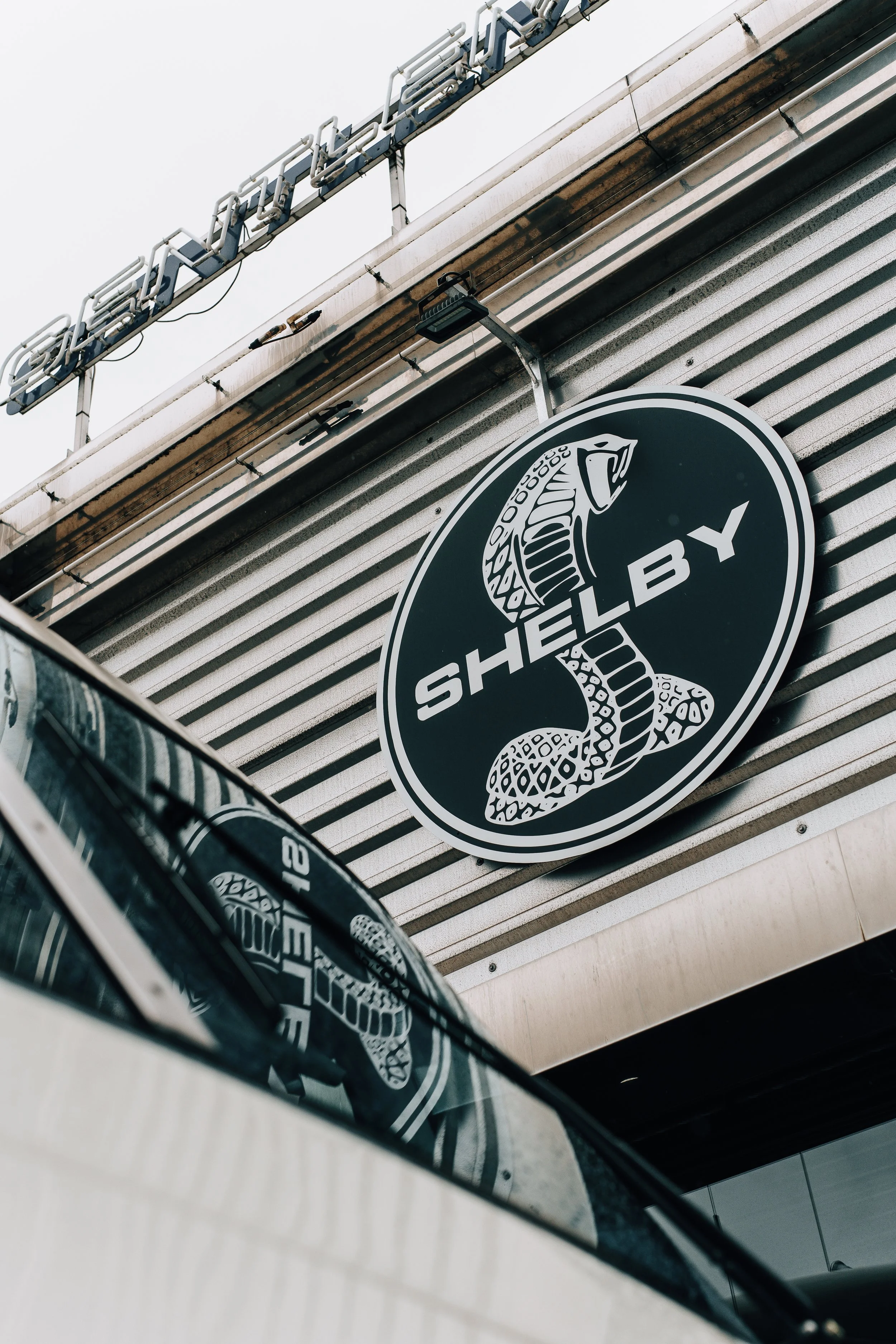 A black circular sign with a white snake illustration and the words 'THE LAB' hanging on a gray wall. Part of a car's side mirror reflects the sign and wall. The background features a branch of a sign with metal piping and a gray sky.