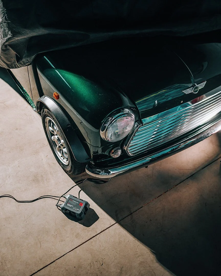 The front part of a vintage black car, possibly a Mini Cooper, parked in a garage. There is an electric device on the floor connected to the car with a cord.