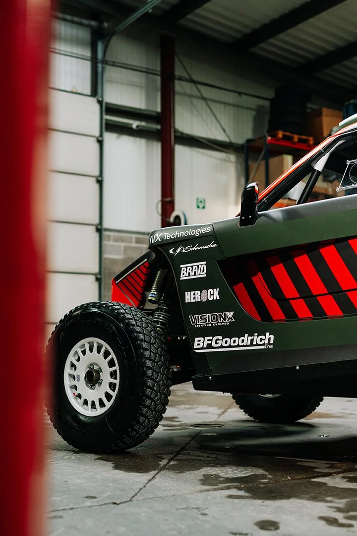 Close-up of a race car with sponsor logos on its side, parked inside a garage or workshop with a concrete floor and industrial walls.