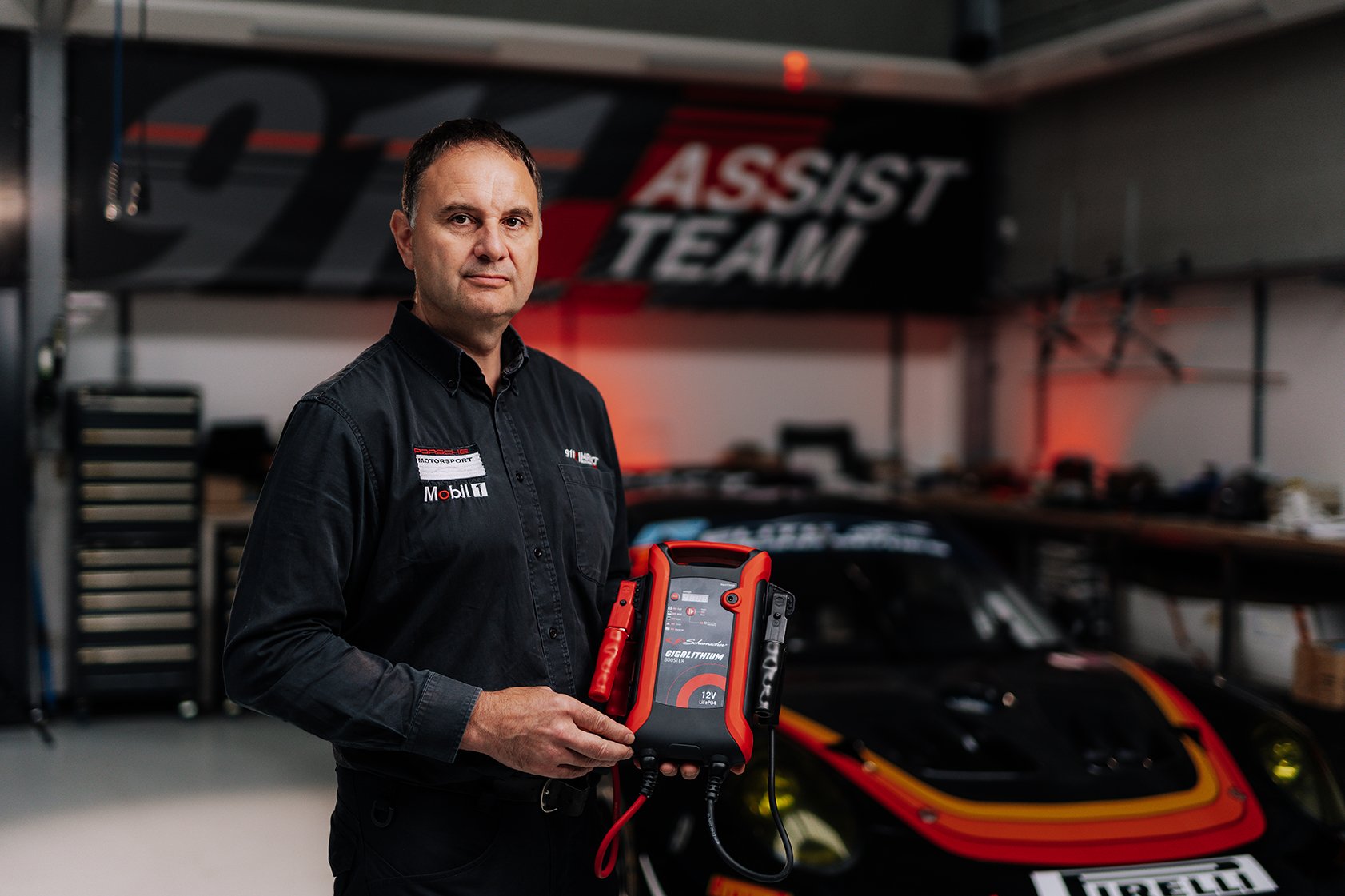 A man in a black racing team shirt holding a 12-volt battery jump starter in a garage with a race car in the background.