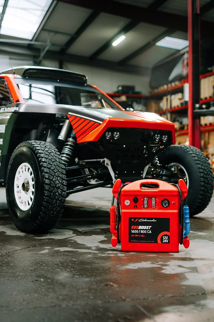 An off-road racing vehicle parked inside a workshop next to a red jumper starter box.