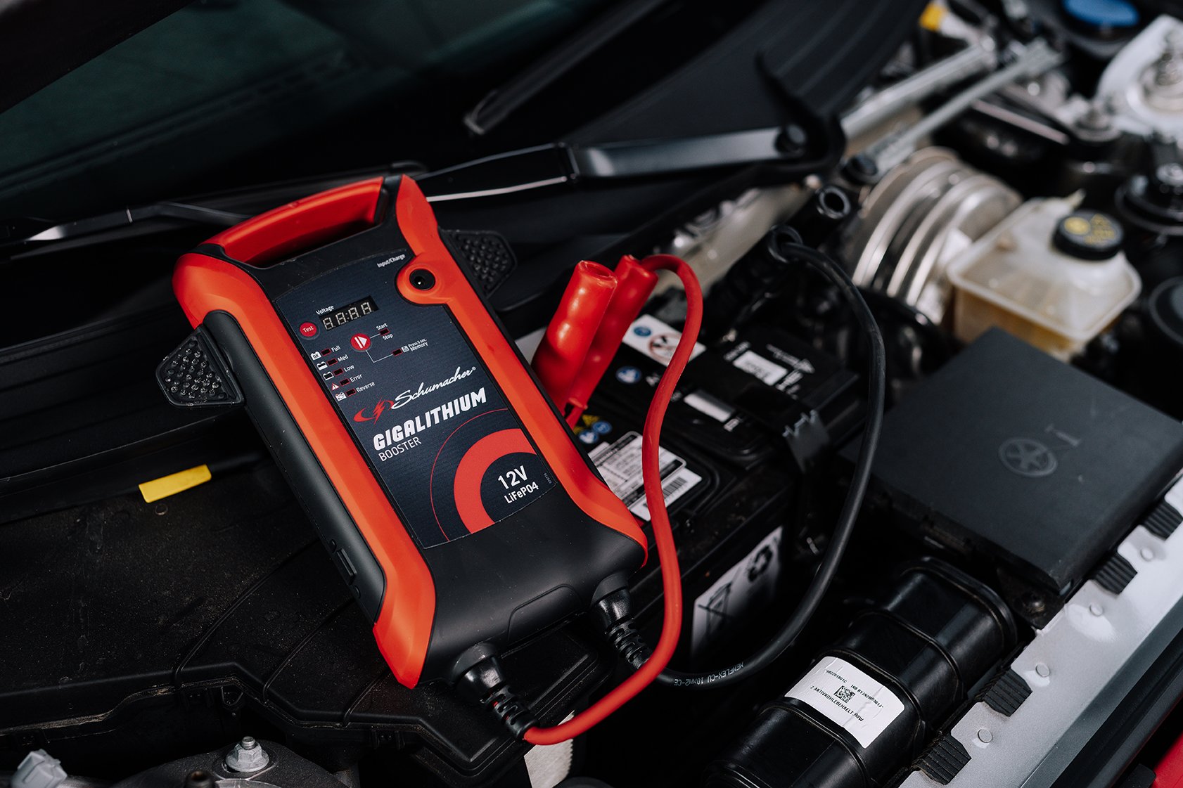 Car battery being tested with a Schumacher GigaLithium booster and multimeter inside the engine bay.
