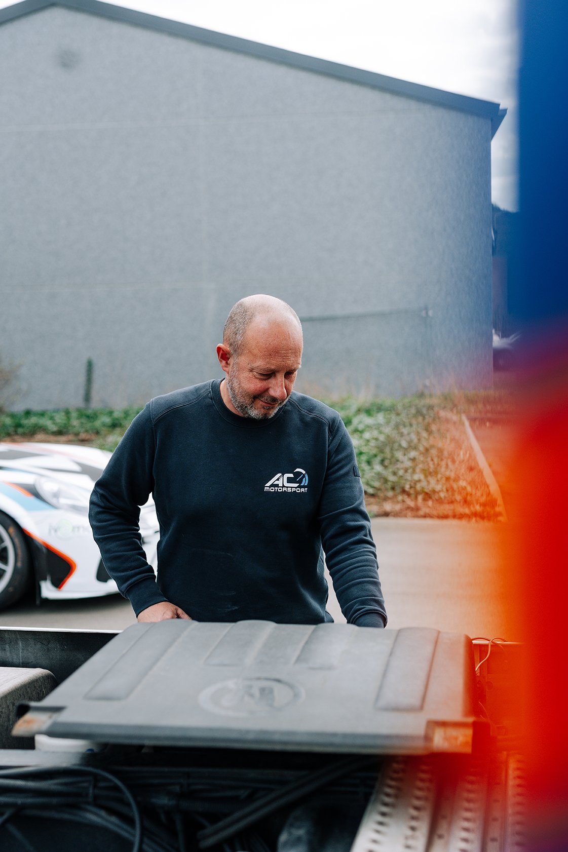 A man with a shaved head and beard wearing a black sweatshirt with 'AC Motorsport' logo, standing outdoors near a race car, with parking lot and building in the background.
